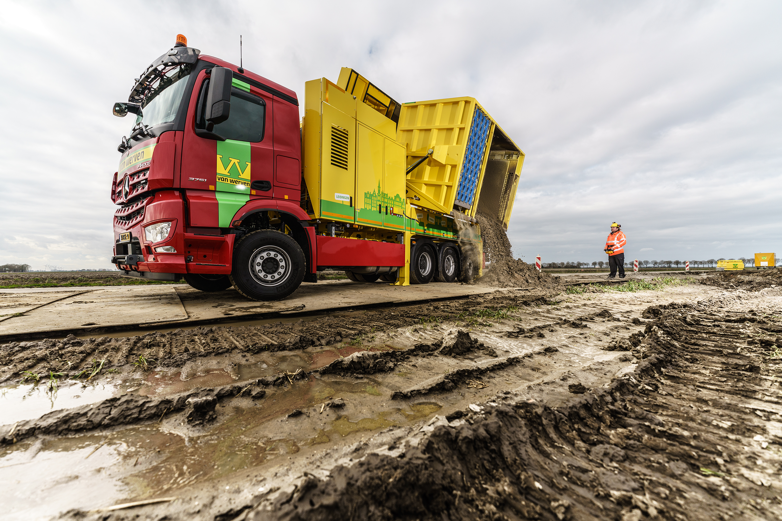 De rode vrachtwagen met geel-groene zuigopbouw lost gezogen grond op het land, onder toezicht van een medewerker