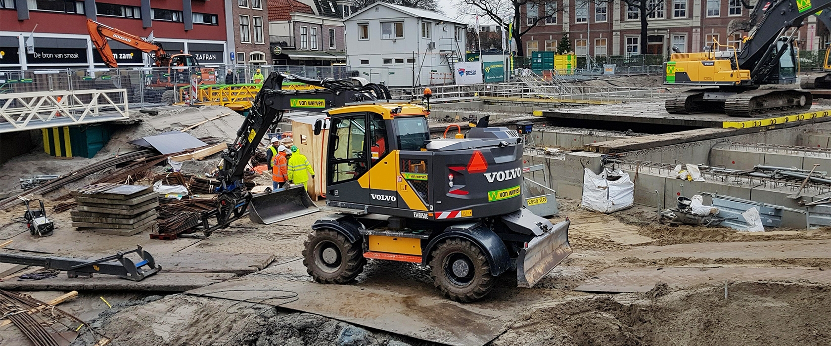 Een Volvo-mobiele kraan van Van Werven met schuifblad op houten rijplaten langs het werkvak, waar grondwerkers overleggen