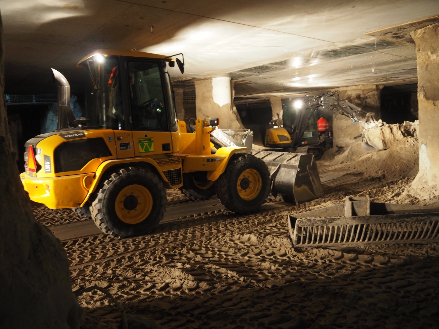 Een compacte Volvo-wiellader van Van Werven rijdt met zand door de lage kelder, een minigraver met werklampen op de achtergrond