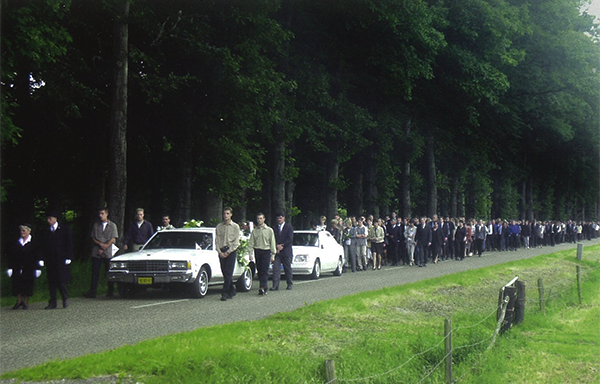 De uitvaartstoet in 2001: twee witte volgauto's en een lange stoet mensen te voet over een bomenlaan