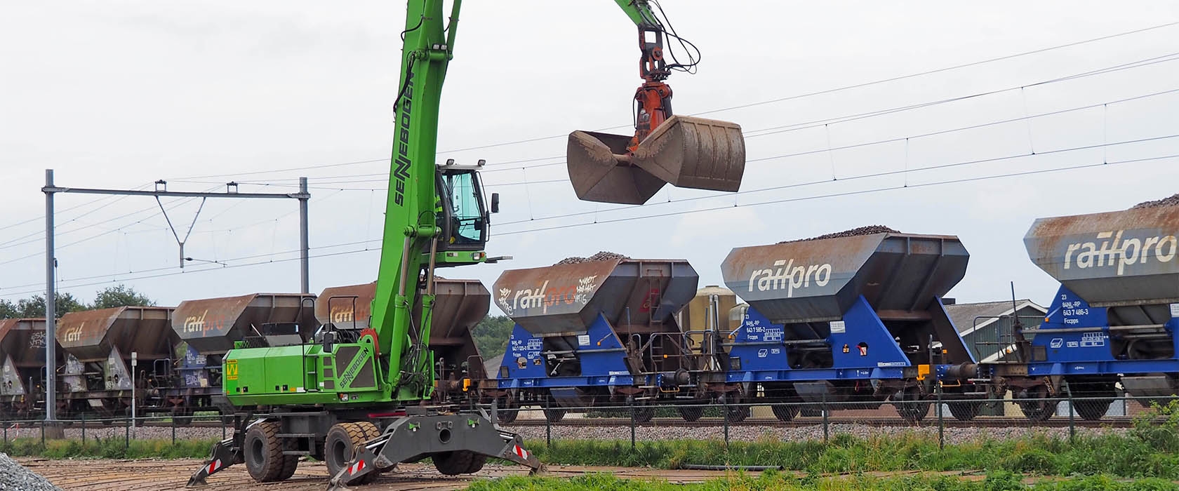 Een groene Sennebogen-overslagkraan tilt een knijperbak met ballast boven een rij blauwe Railpro-goederenwagons