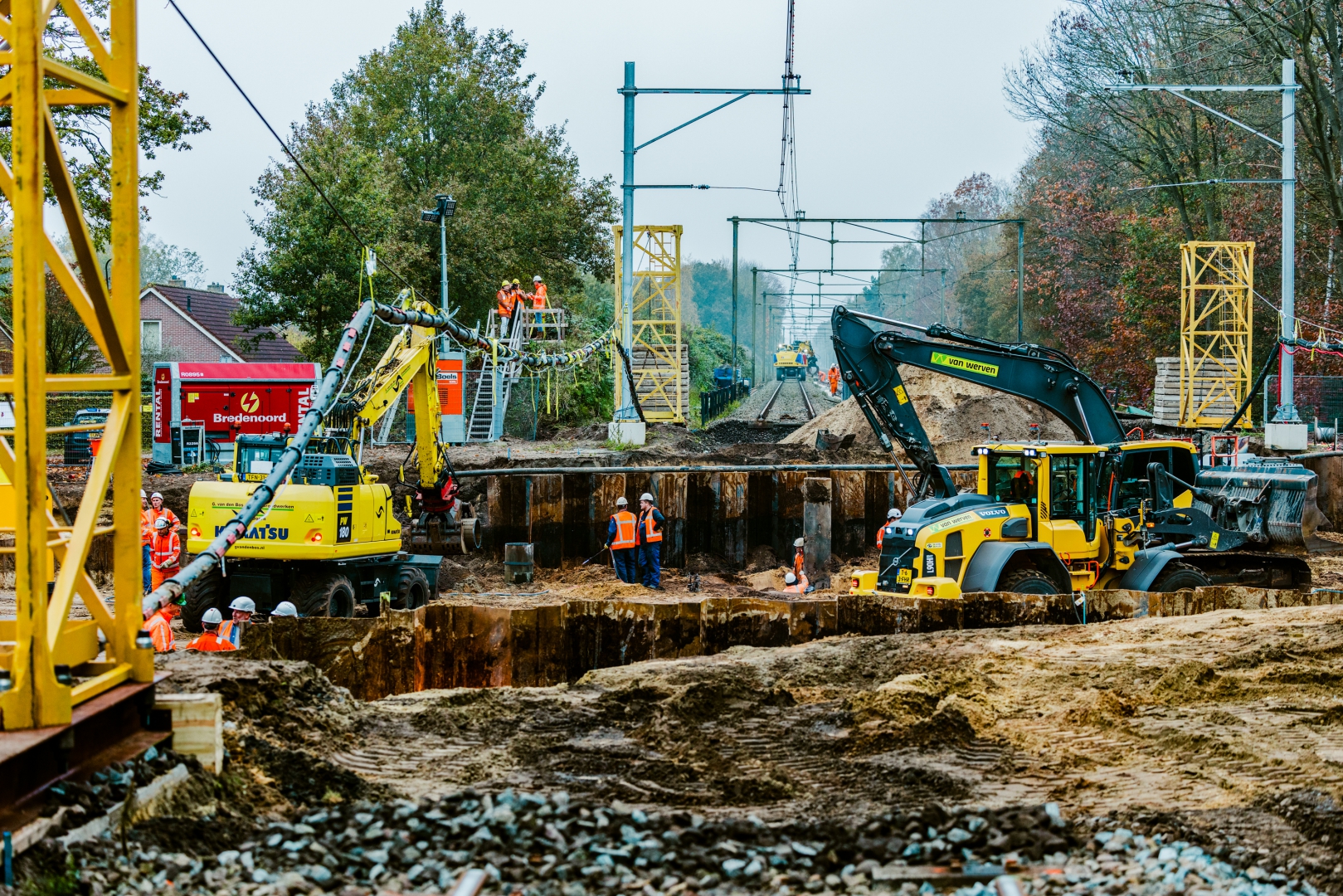 Spoorbouwplaats: een Komatsu-kraan en een Volvo wiellader van Van Werven werken in een bouwput met damwanden onder de bovenleiding, met werklieden in oranje