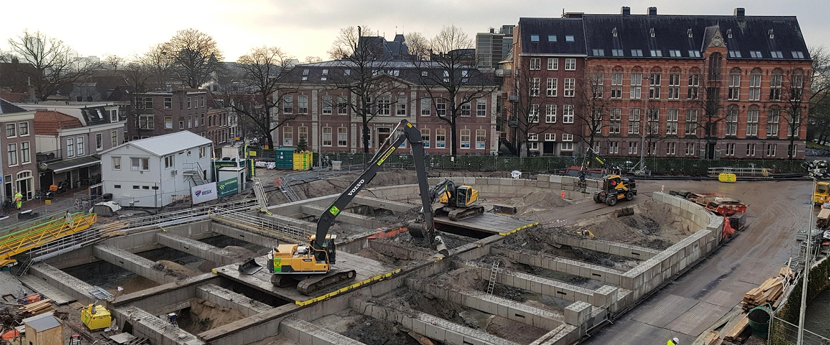 De bouwkuip in de winterschemering, met kranen op het werkdek en de monumentale schoolgebouwen langs de rand