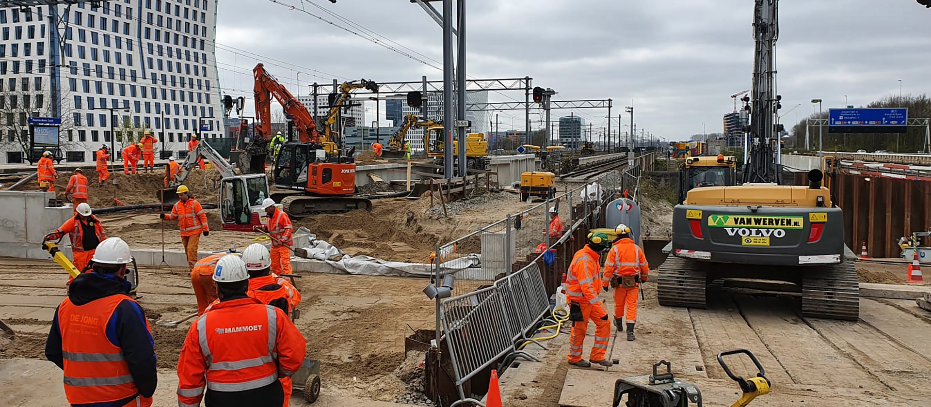 Overzicht van het spoorwerkterrein bij station Amsterdam Zuid: tientallen collega's in oranje, graafmachines en een heistelling tussen de bovenleidingportalen