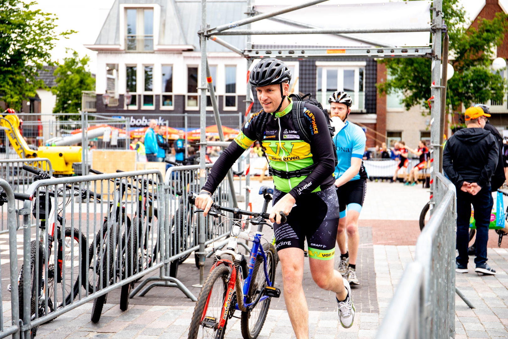 Een deelnemer in Van Werven-wielershirt duwt zijn mountainbike tussen de dranghekken door, een tweede renner erachter