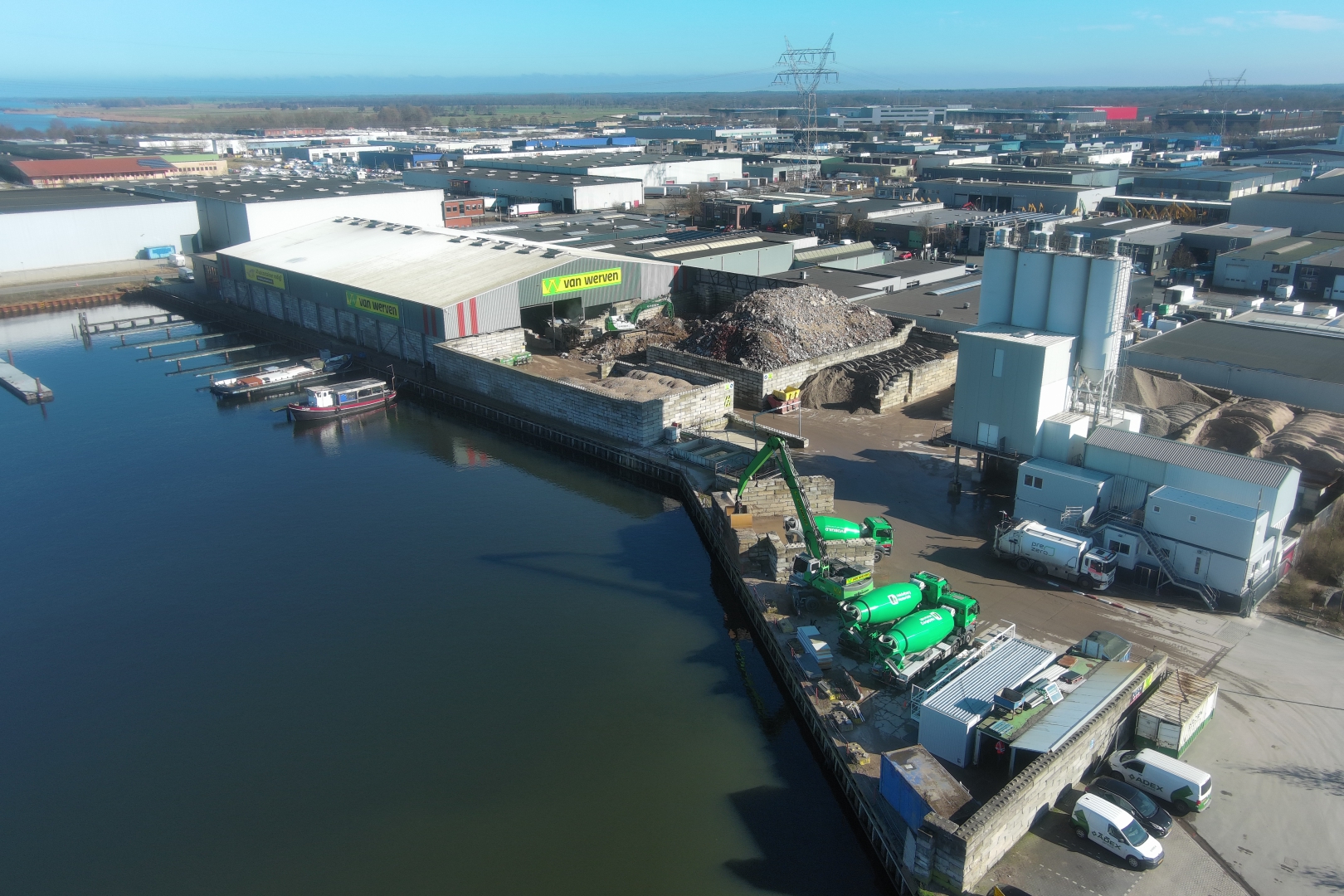 Luchtfoto van Recycling Harderwijk aan het water, met de loods, puinbergen, betonsilo's en groene betonmixers