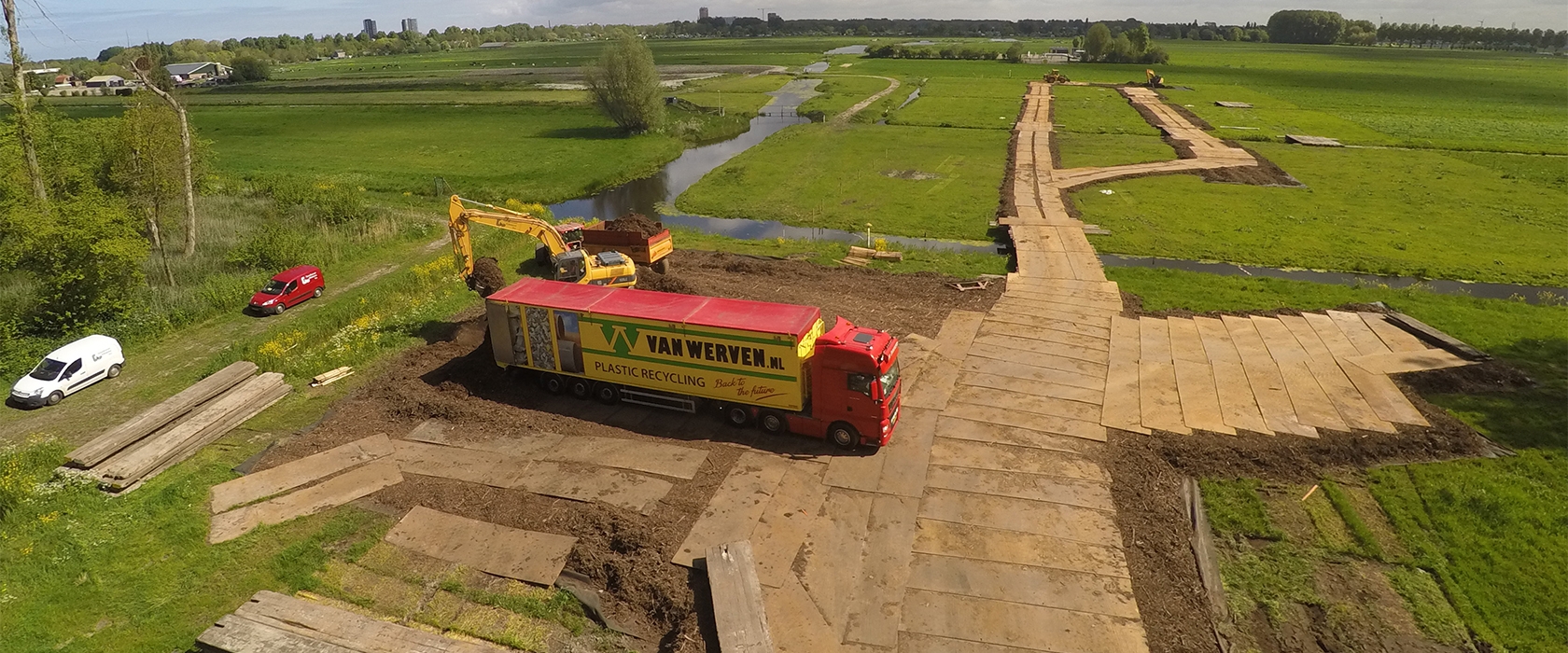 Luchtfoto van een tijdelijke rijbaan van rijbaanhout die door de weilanden bij Leiden loopt, met een vrachtwagen van Van Werven en een graafmachine erop