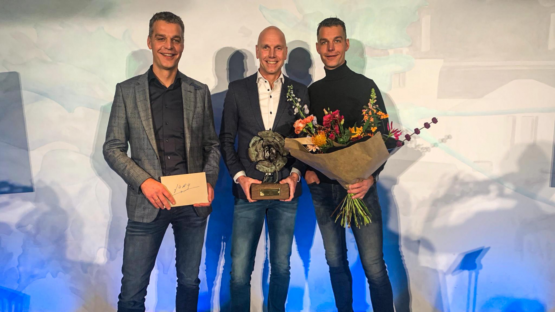 Drie mannen op het podium met de bronzen award, een bloemboeket en het juryrapport