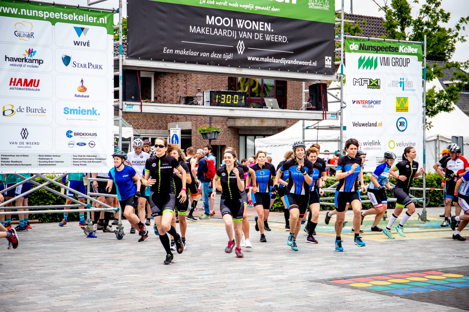Een groep deelnemers sprint weg onder de startboog met sponsordoeken; de klok erboven staat op 1:30:01