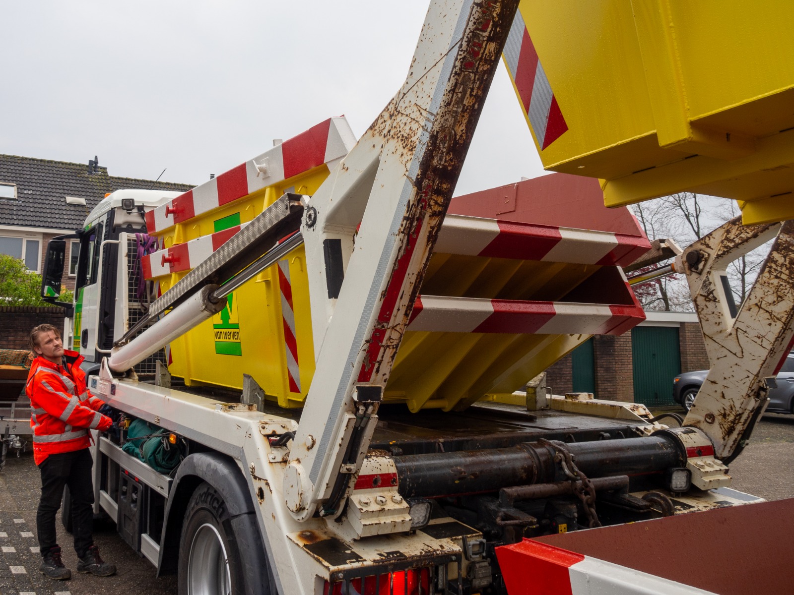 Een medewerker in oranje jas bedient de portaalarmwagen die een gele container optilt in de woonstraat