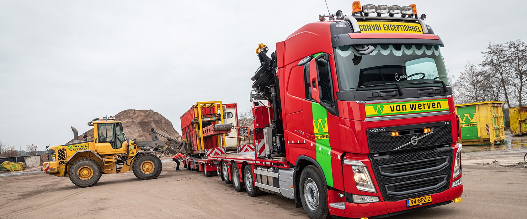 Nieuwe rode Volvo FH-truck van Van Werven met dieplader en autolaadkraan, met een gele Volvo wiellader op de achtergrond