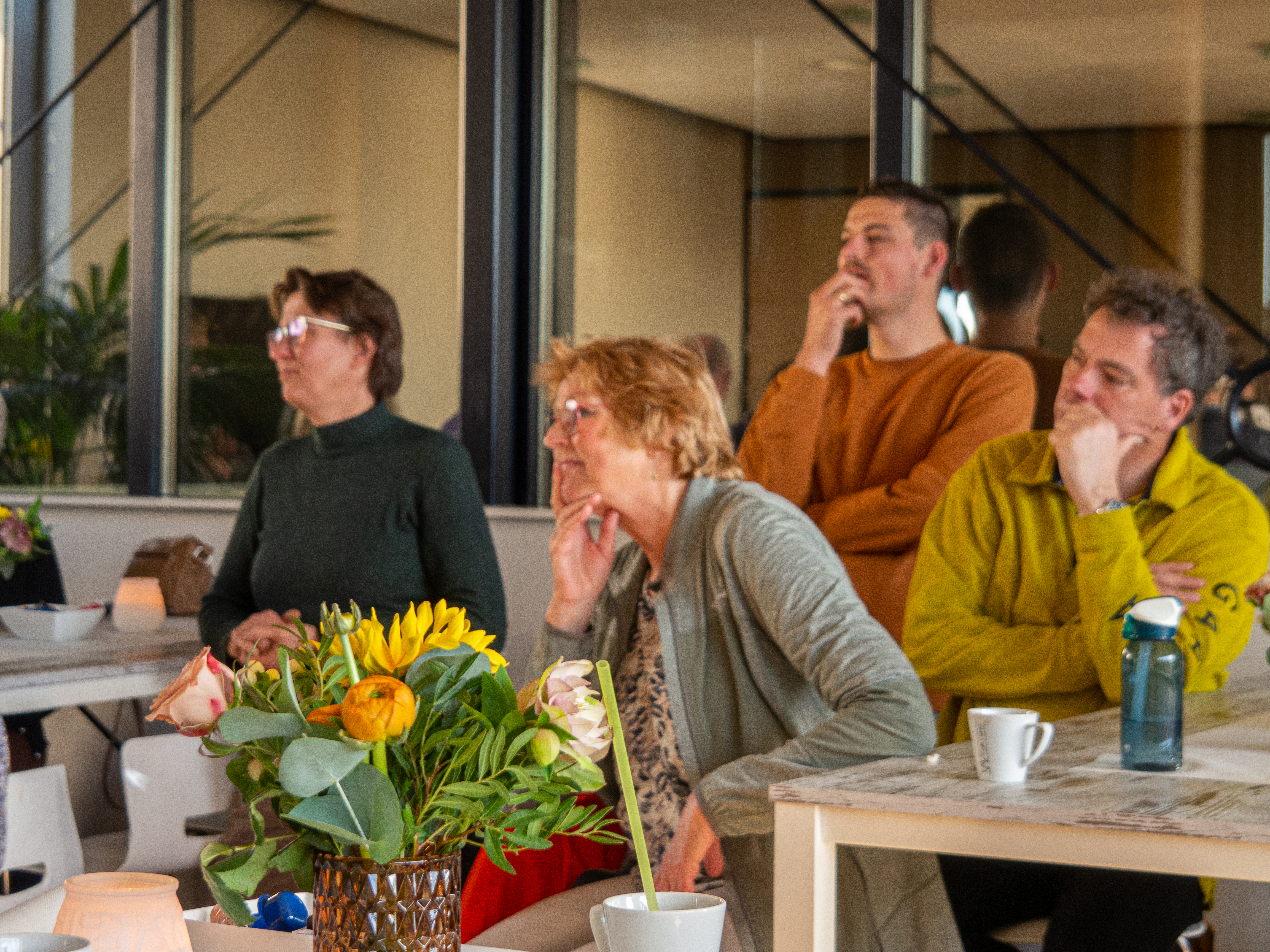 Genodigden luisteren aandachtig aan tafels met bloemen en koffie tijdens de bijeenkomst