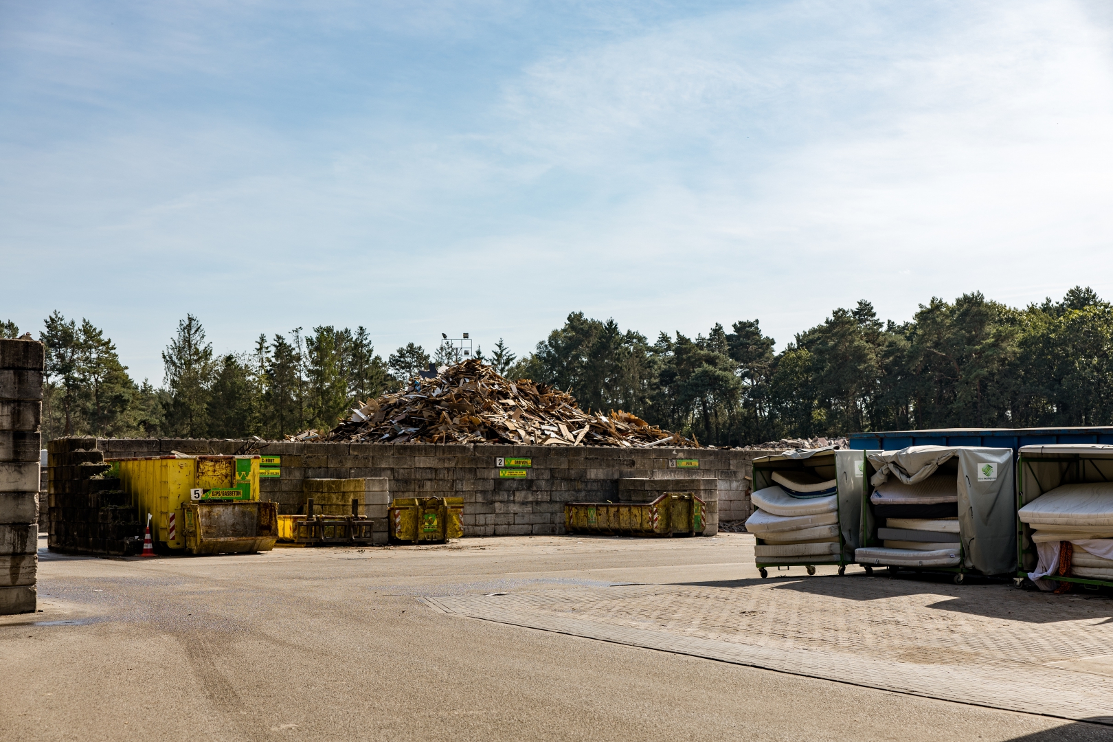 De milieustraat met een berg houtafval achter betonblokken, gele containers en rolcontainers vol matrassen