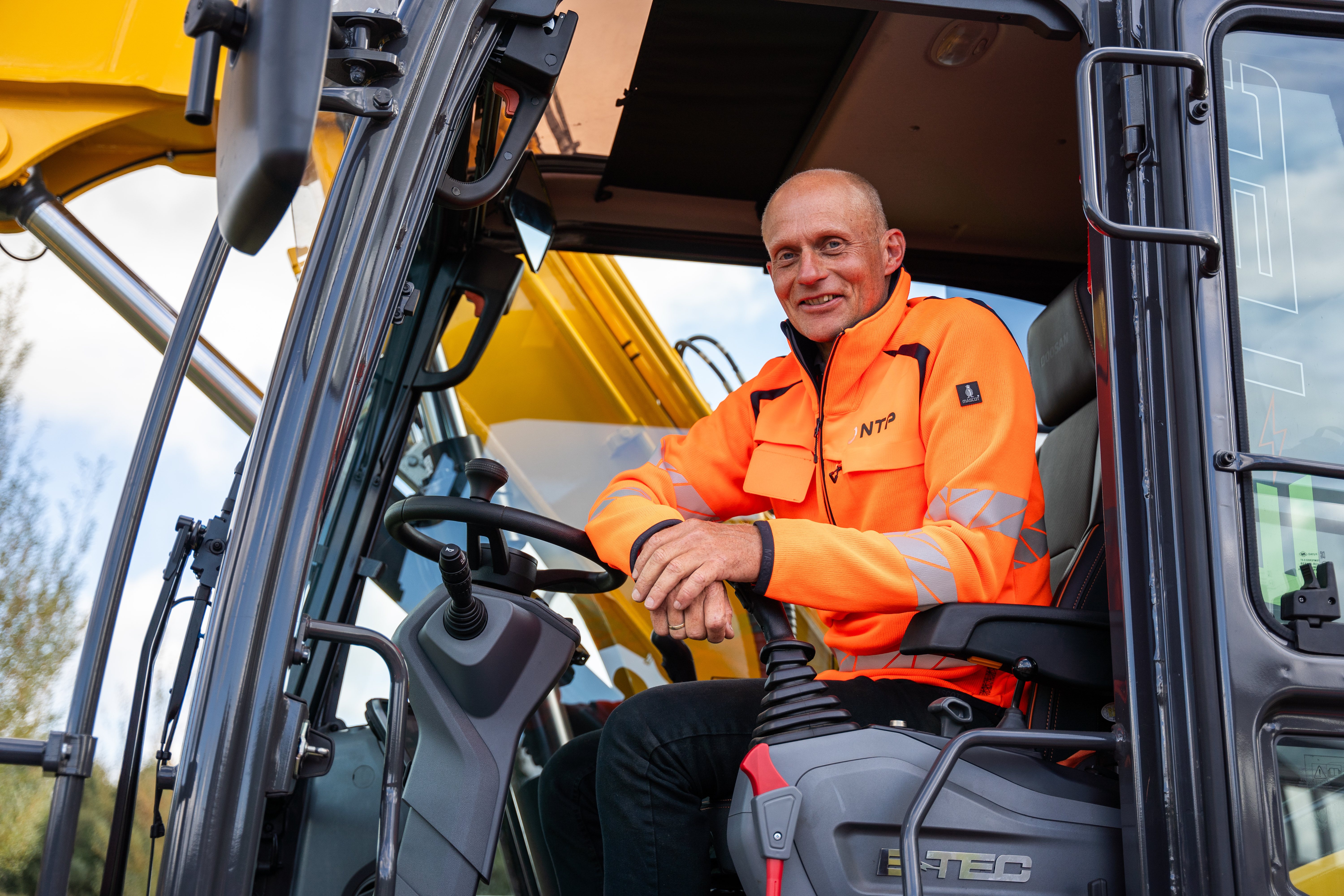 Een machinist in oranje NTP-jas lacht vanuit de cabine van de elektrische E-TEC-kraan