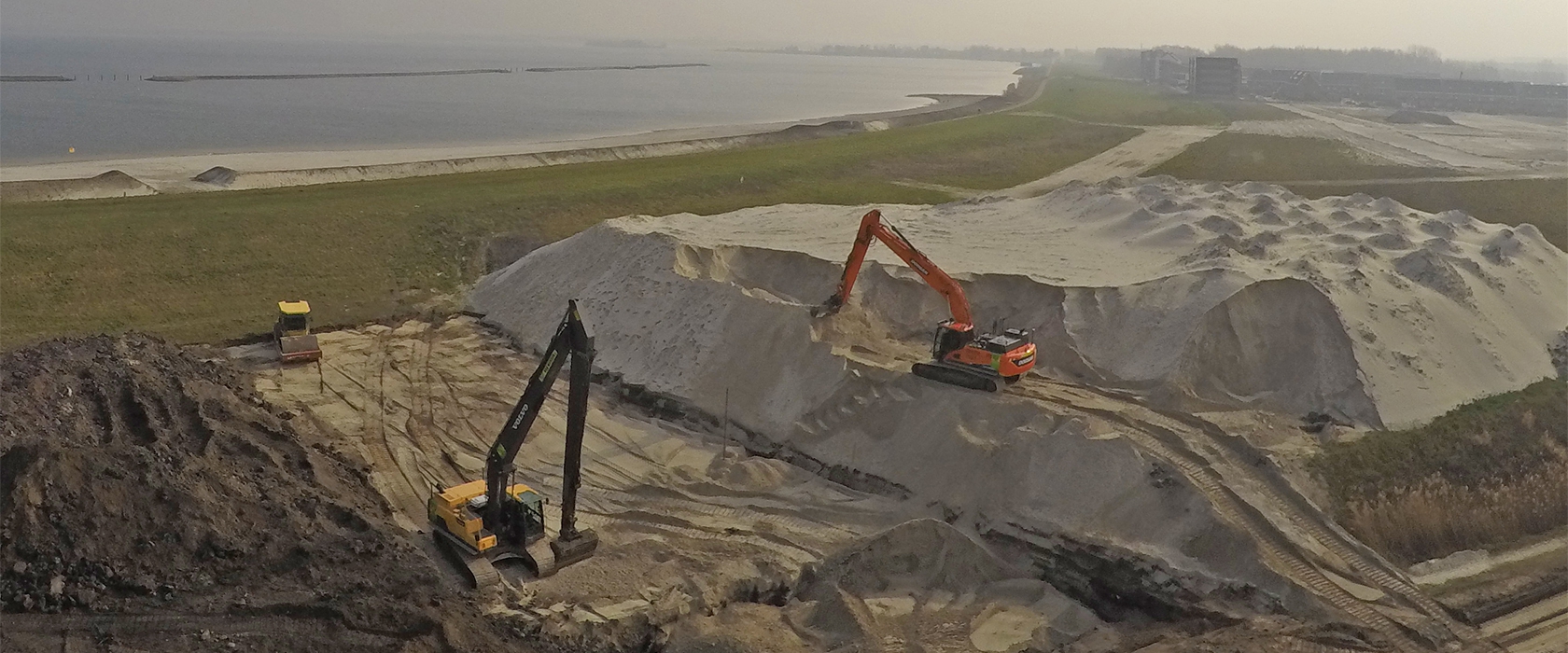 Twee rupskranen met lange giek werken op een groot zanddepot aan het randmeer, naast de groene dijk met appartementen erachter