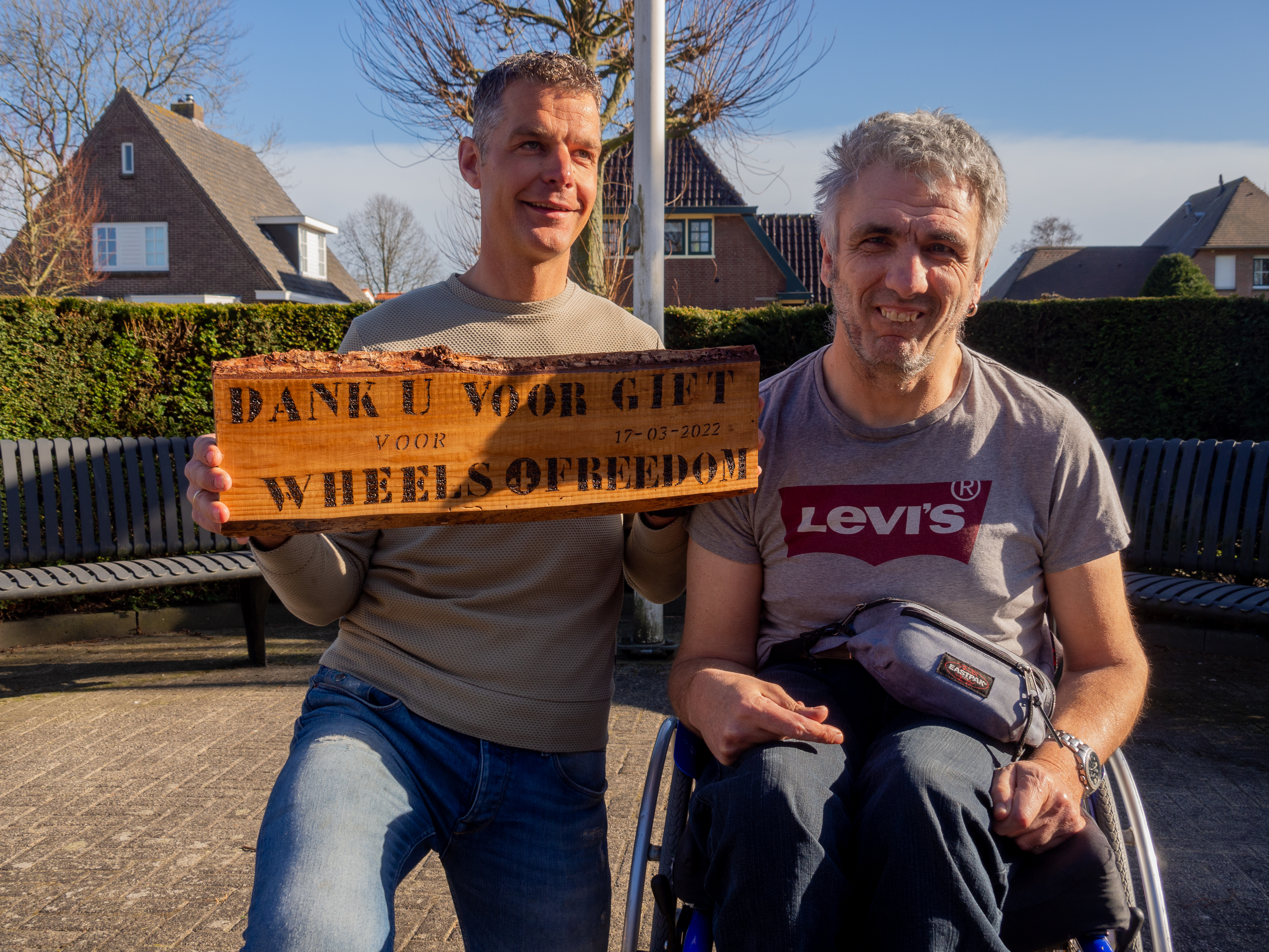 Twee mannen, van wie één in een rolstoel, met een houten bord met de tekst 'Dank u voor gift voor Wheels of Freedom'