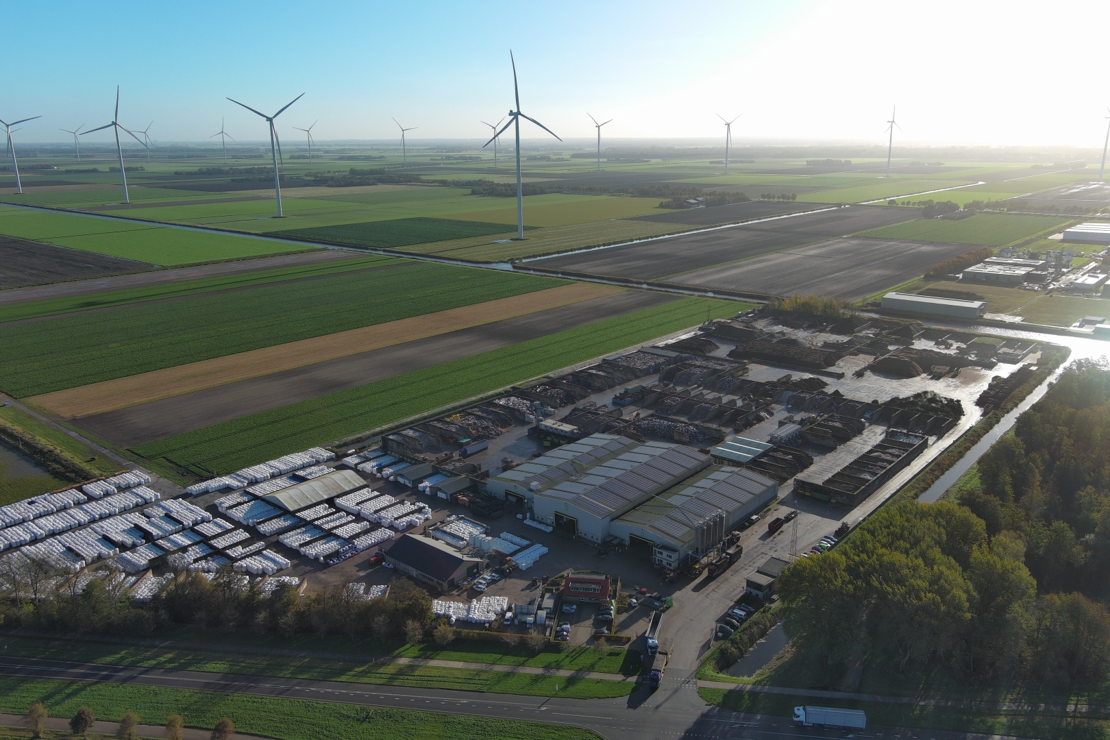 Luchtfoto van de kunststofrecyclinglocatie met rijen witte big bags en loodsen, omringd door akkers en windmolens