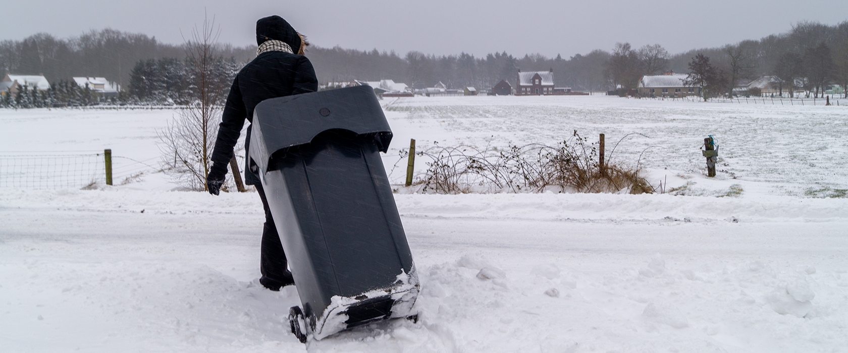 Iemand in winterjas trekt een zwarte kliko door de sneeuw langs een besneeuwde weg