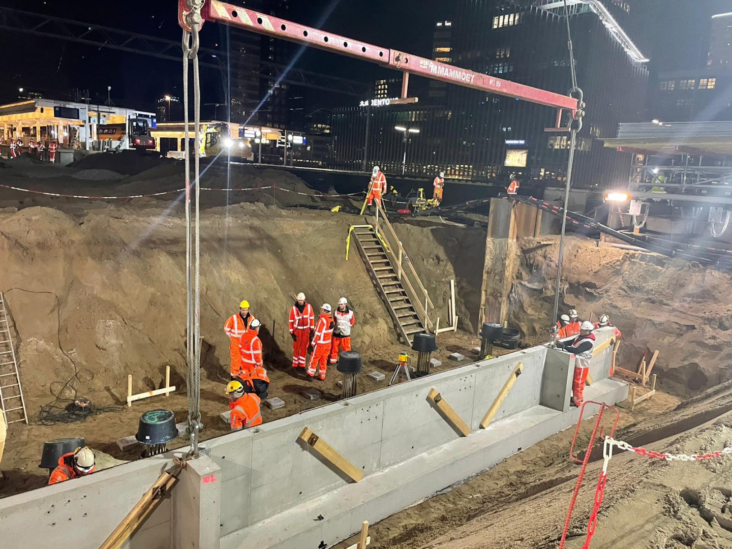 Nachtwerk: aan het hijsjuk van Mammoet hangt een betonnen wandelement boven de put, met het station op de achtergrond