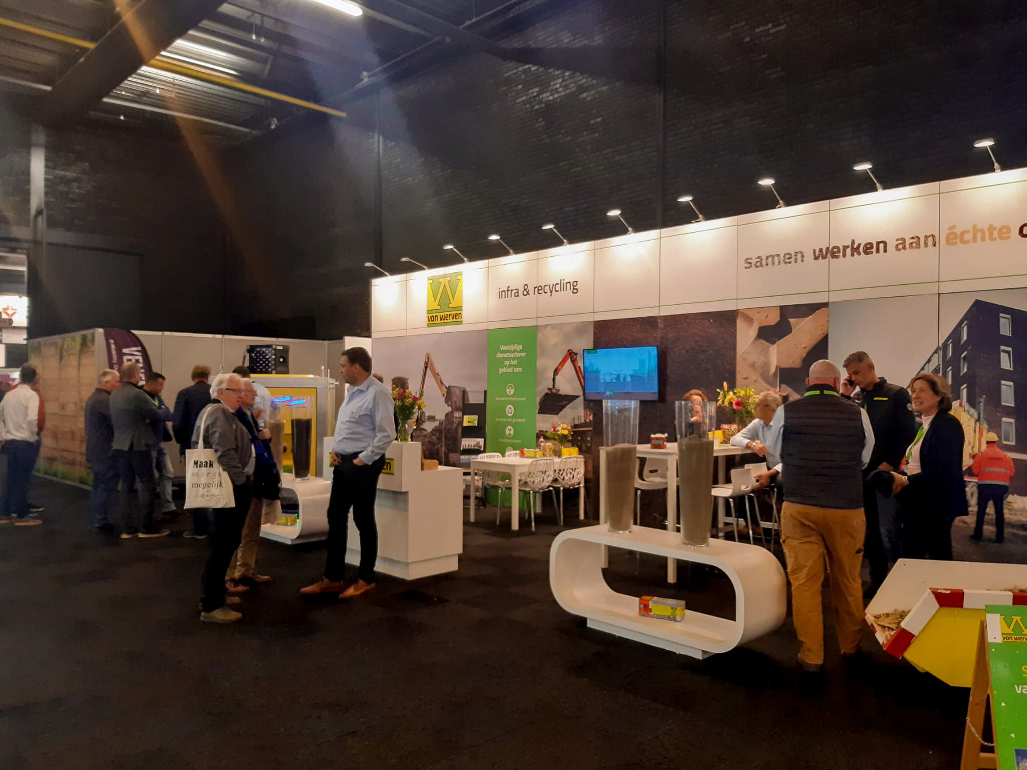 Bezoekers in gesprek bij de Van Werven-stand op de Infra Relatiedagen, met glazen kolommen vol grondmonsters en een tv-scherm