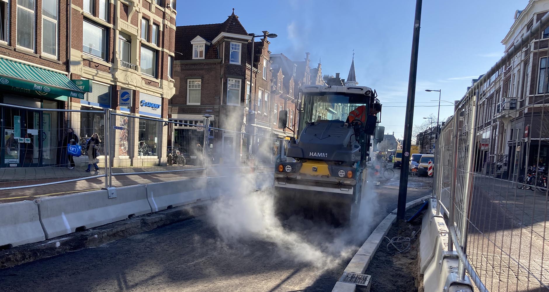 Een dampende asfaltwals tussen de betonnen barriers in de winkelstraat, met winkelend publiek achter het bouwhek