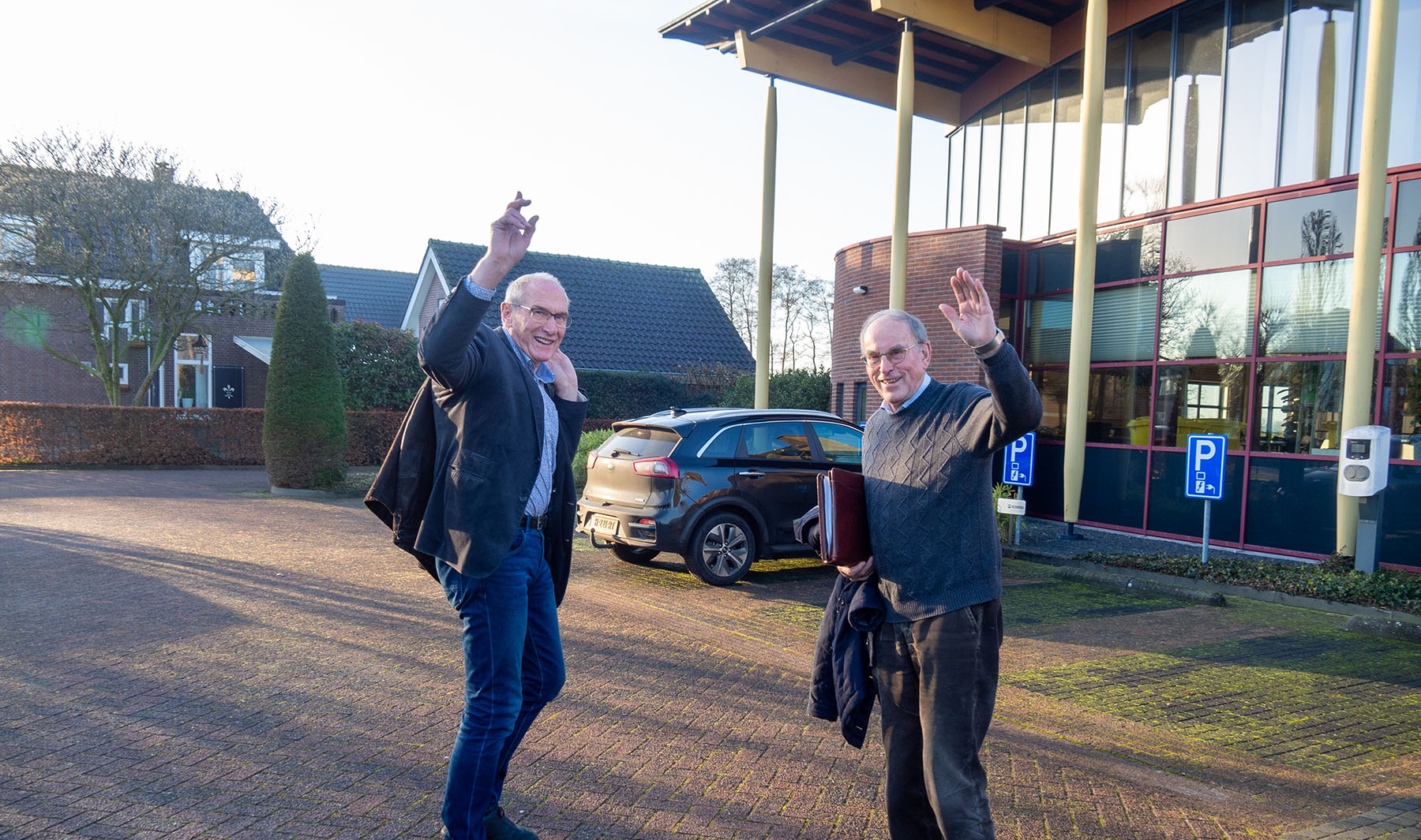 Twee zwaaiende mannen op het parkeerterrein voor het kantoor, een van hen met een map onder de arm