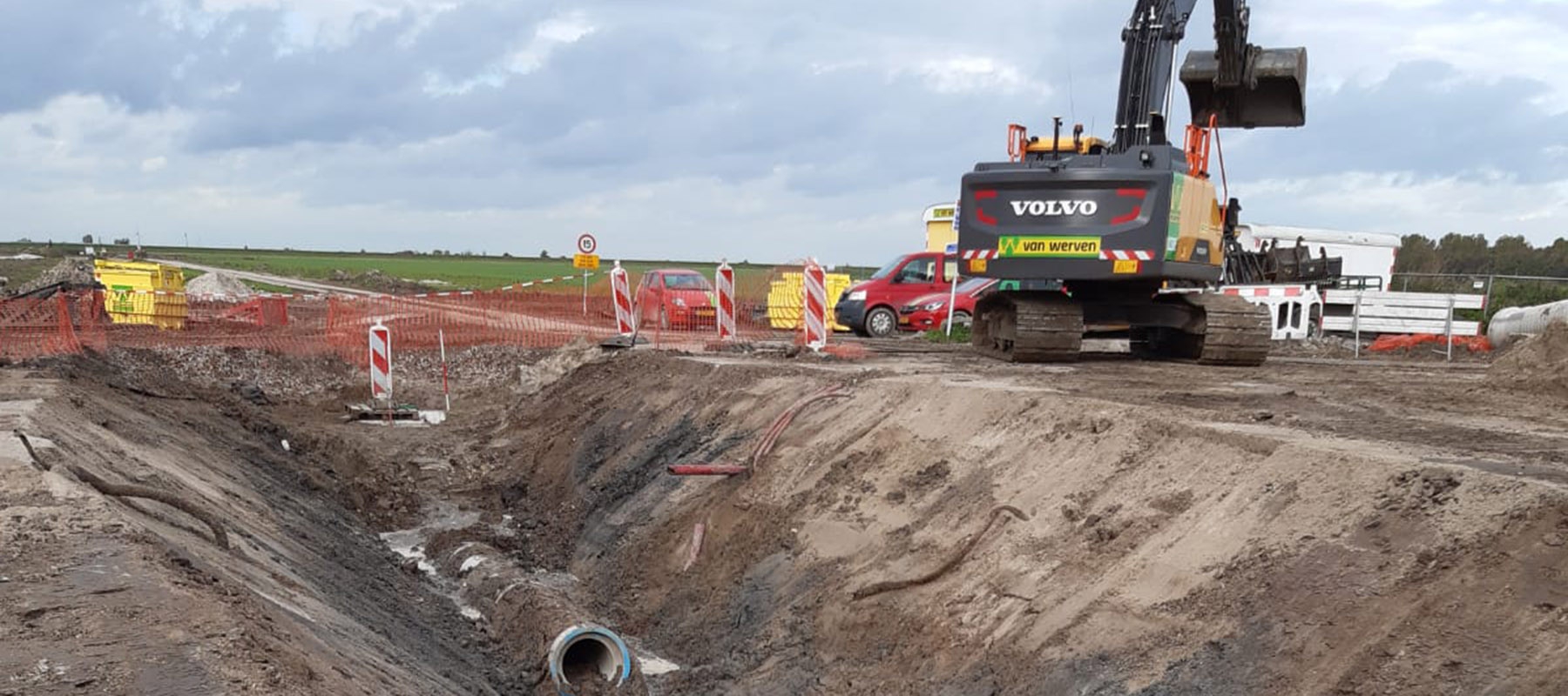 Een Volvo-rupskraan van Van Werven bij een diepe sleuf met een rioolbuis, achter de rood-witte afzetting langs de weg