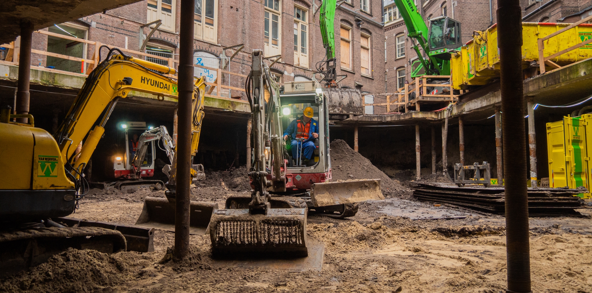 Minigravers ontgraven de kelder onder het monumentale universiteitsgebouw, dat op stalen buispalen rust; bovenlangs reikt de arm van een groene overslagkraan