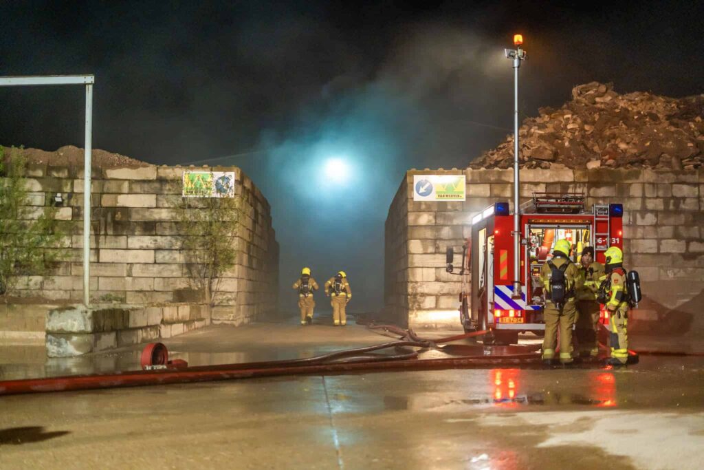 Brandweerlieden met slangen bij een brandweerwagen tussen betonblokwanden in de nacht, met rook boven het terrein