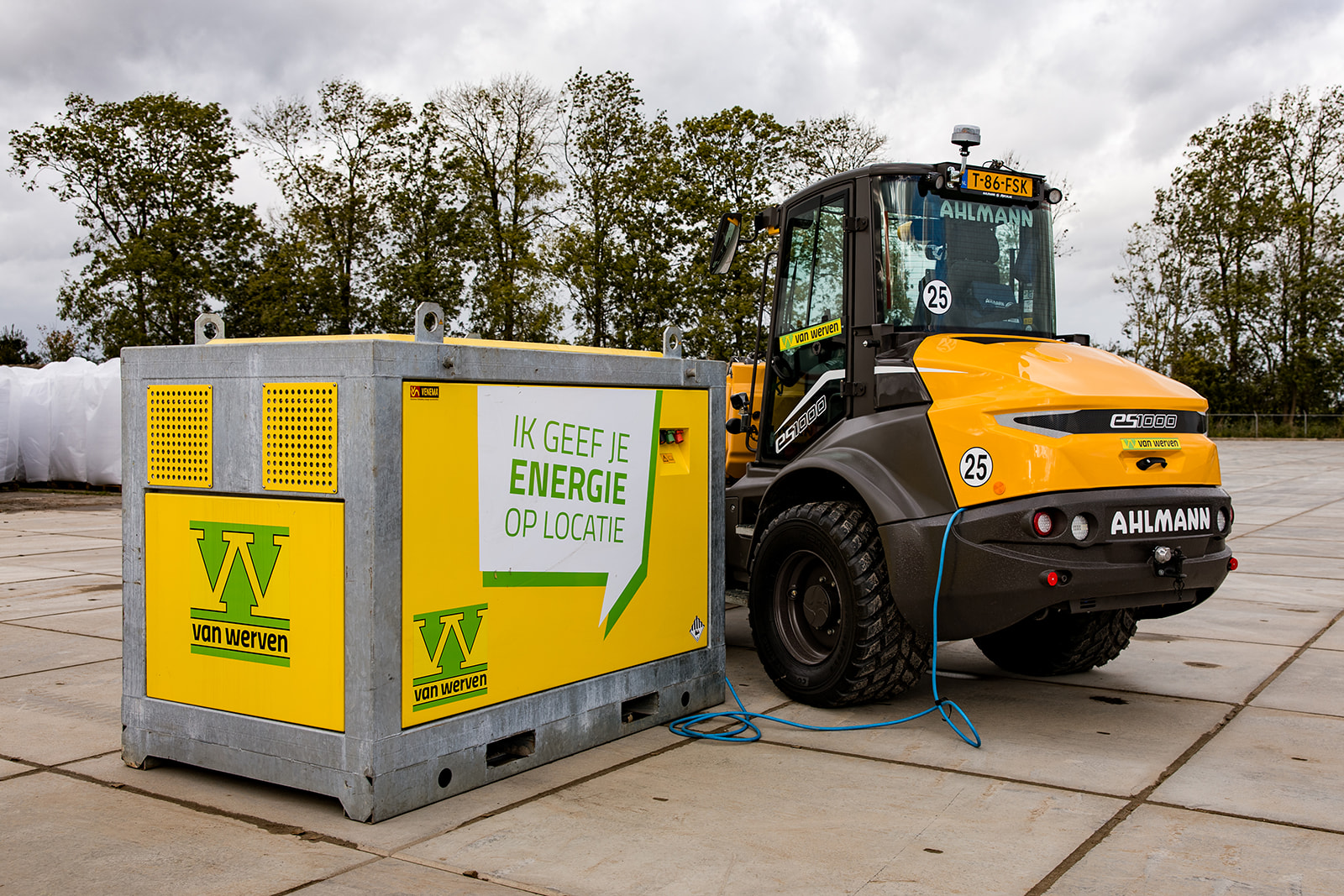 Gele Van Werven-accubox met de tekst 'Ik geef je energie op locatie', via een blauwe kabel verbonden met een elektrische Ahlmann eS1000 shovel