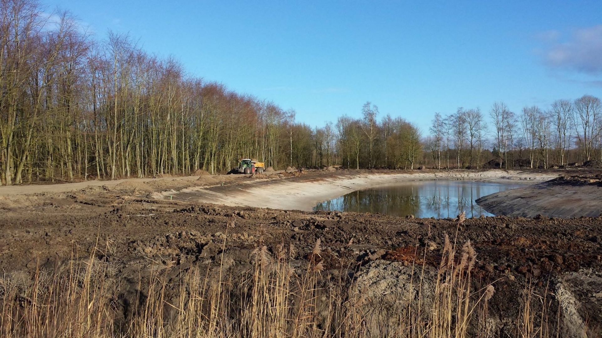 De nieuw gegraven waterplas met licht zandstrand, met een trekker met dumperwagen op het pad langs de bosrand