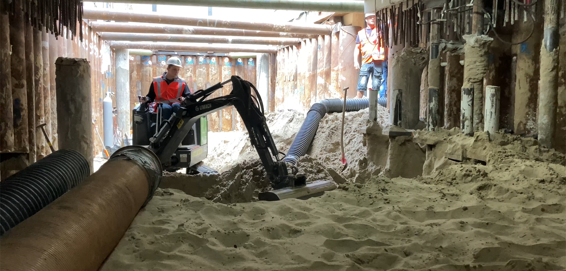 Minigraver in een diepe bouwput tussen damwanden; een zuigslang voert het zand af