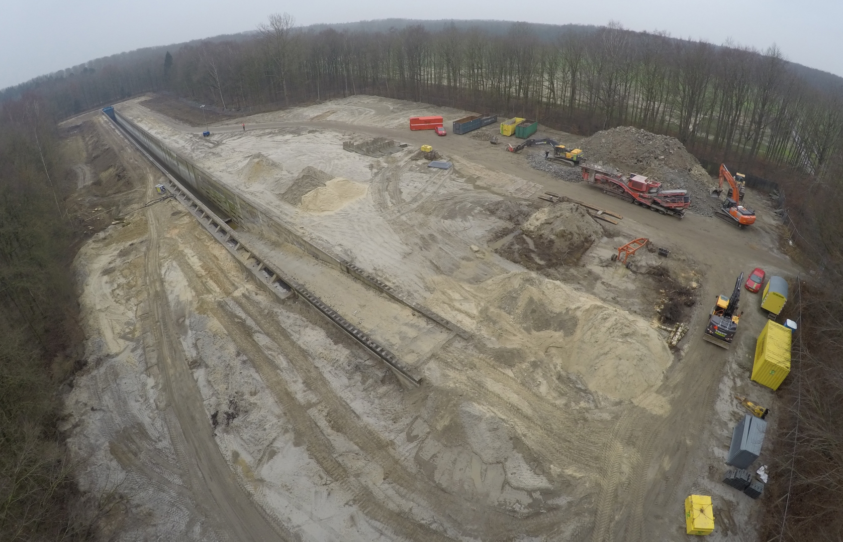 Luchtfoto van het uitgegraven werkterrein naast de betonnen goot, met zandhopen, een puinbreker en containers