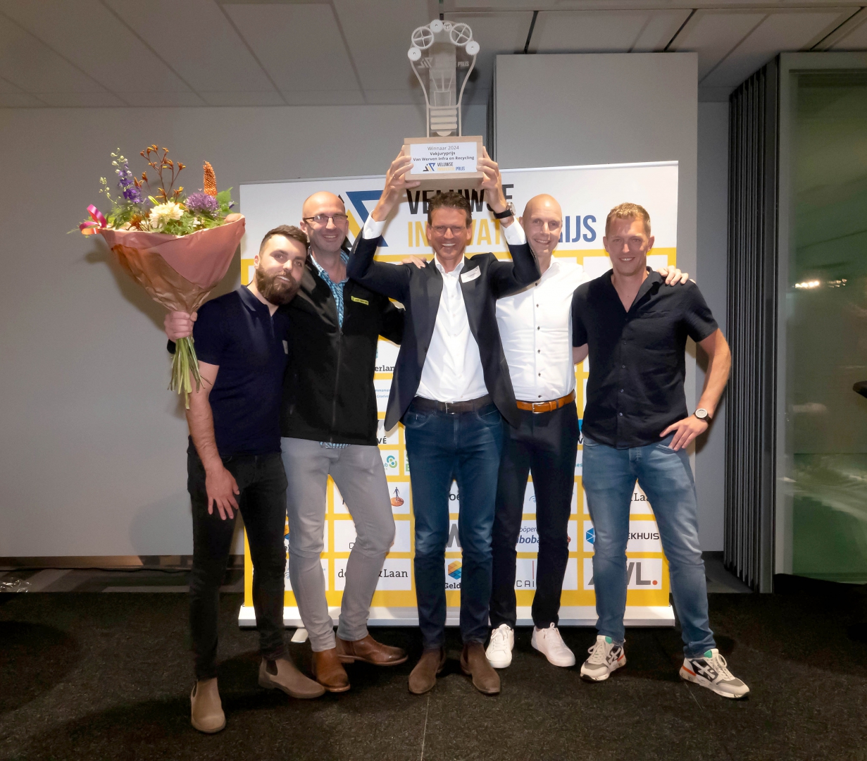 Vijf mannen vieren de winst: de middelste houdt de trofee van de Veluwse Innovatieprijs 2024 boven zijn hoofd, links een bloemboeket