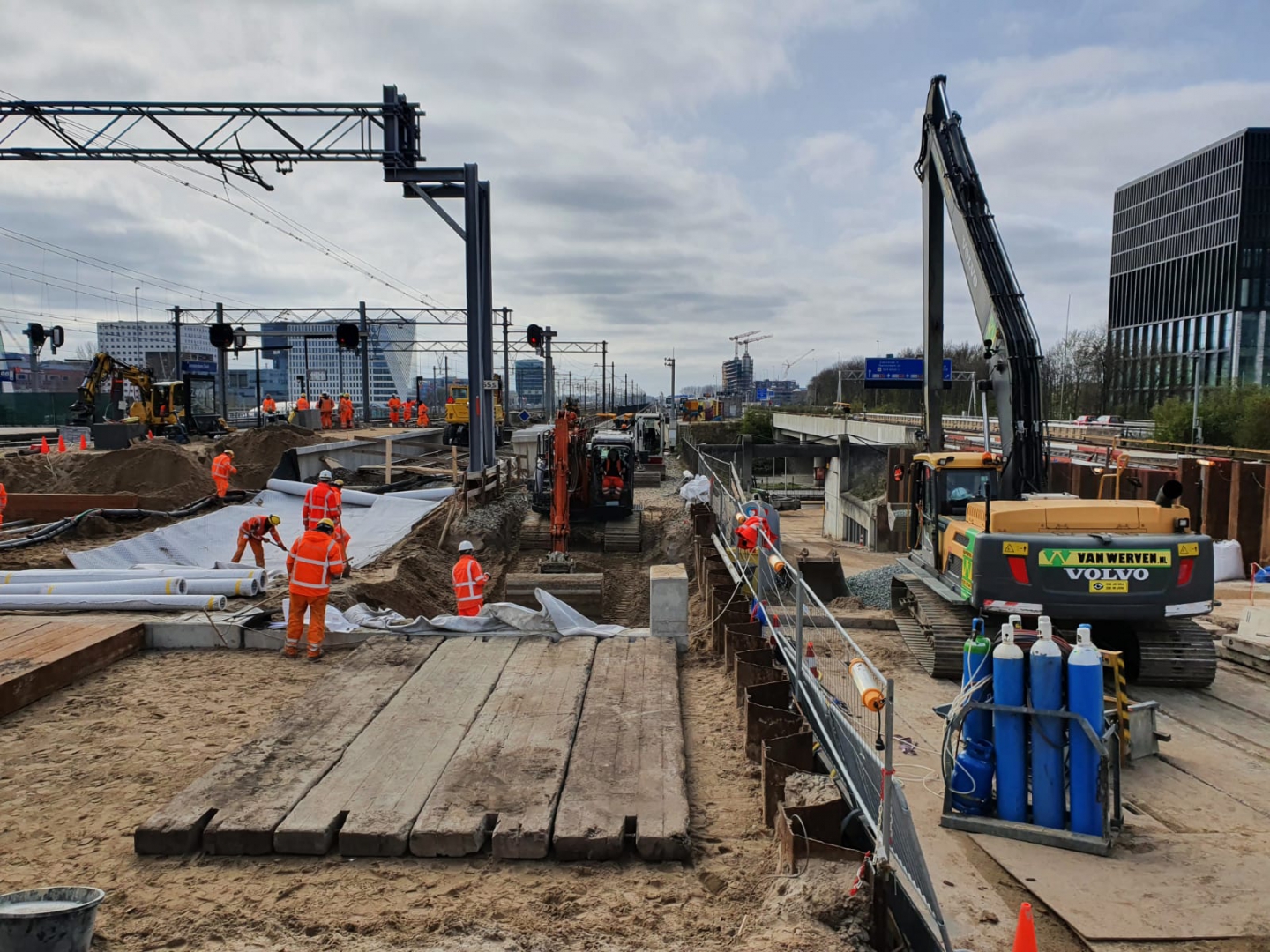 Medewerkers leggen wit doek in de ontgraving langs het spoor; rechts een rupskraan met lange giek, vooraan houten rijplaten