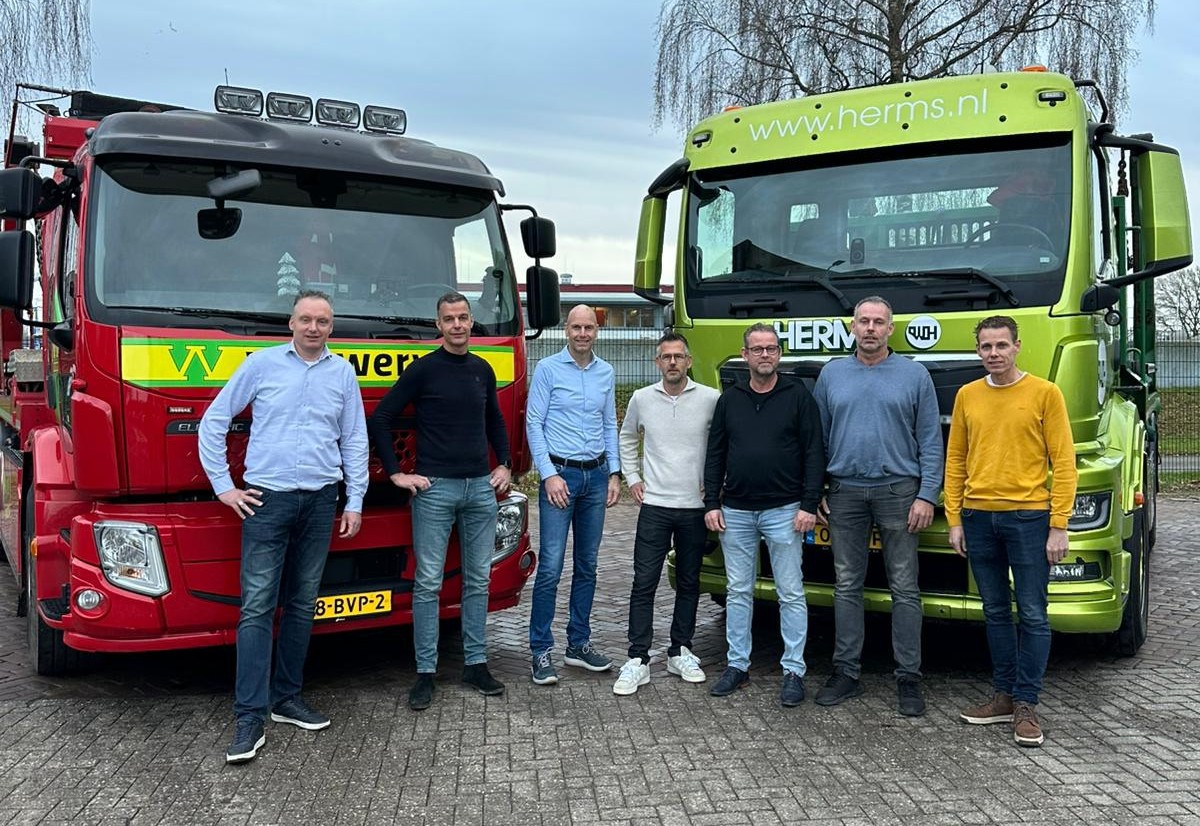 Zeven mannen poseren tussen een rode elektrische vrachtwagen van Van Werven en een limoengroene vrachtwagen van Herms