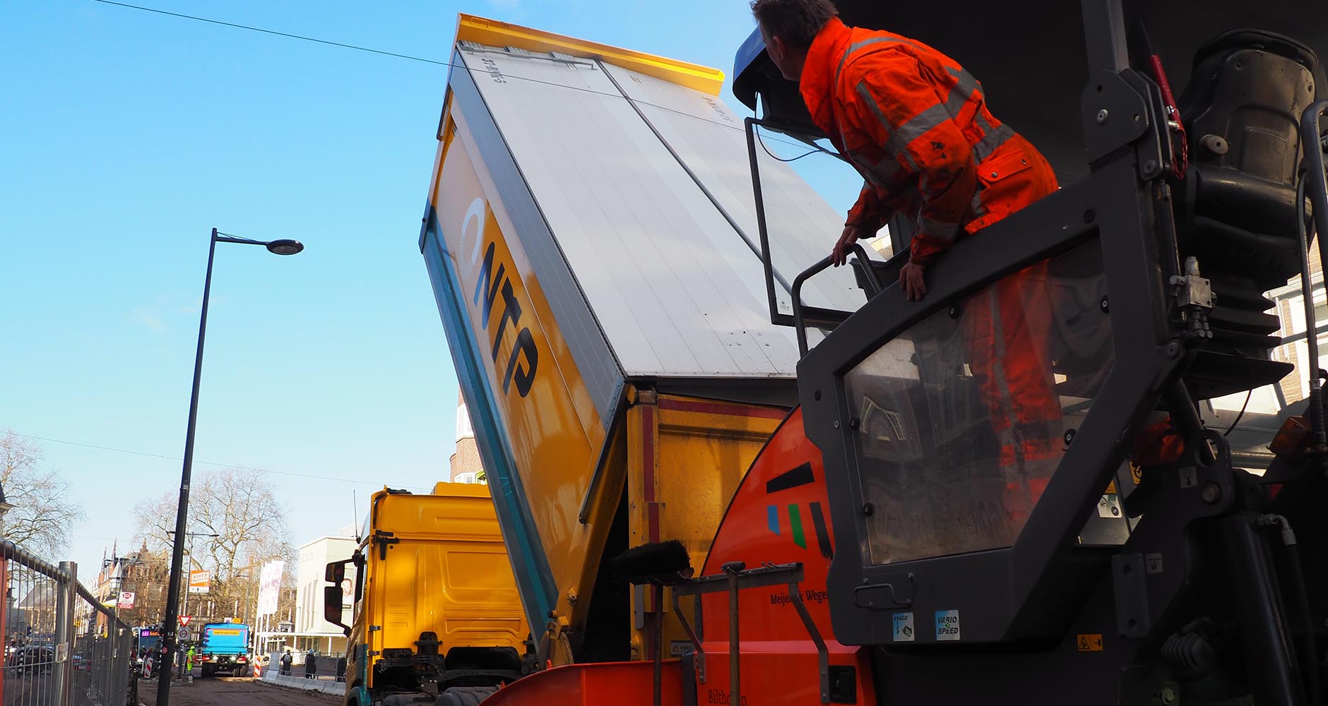 Een kipper van NTP stort asfalt in de asfalteermachine, terwijl een man in oranje vanaf de machine meekijkt