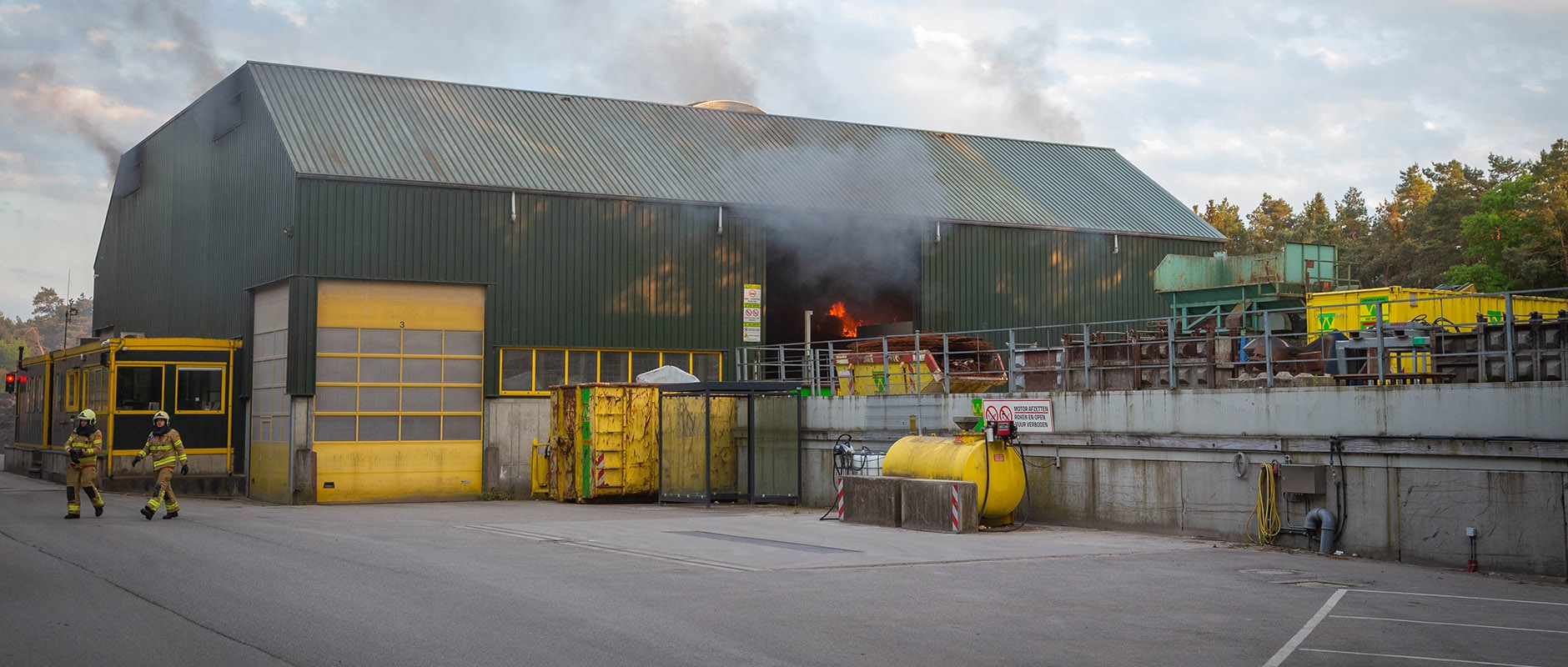Rook stijgt op uit de groene recyclinghal in Oldebroek terwijl twee brandweerlieden over het terrein lopen