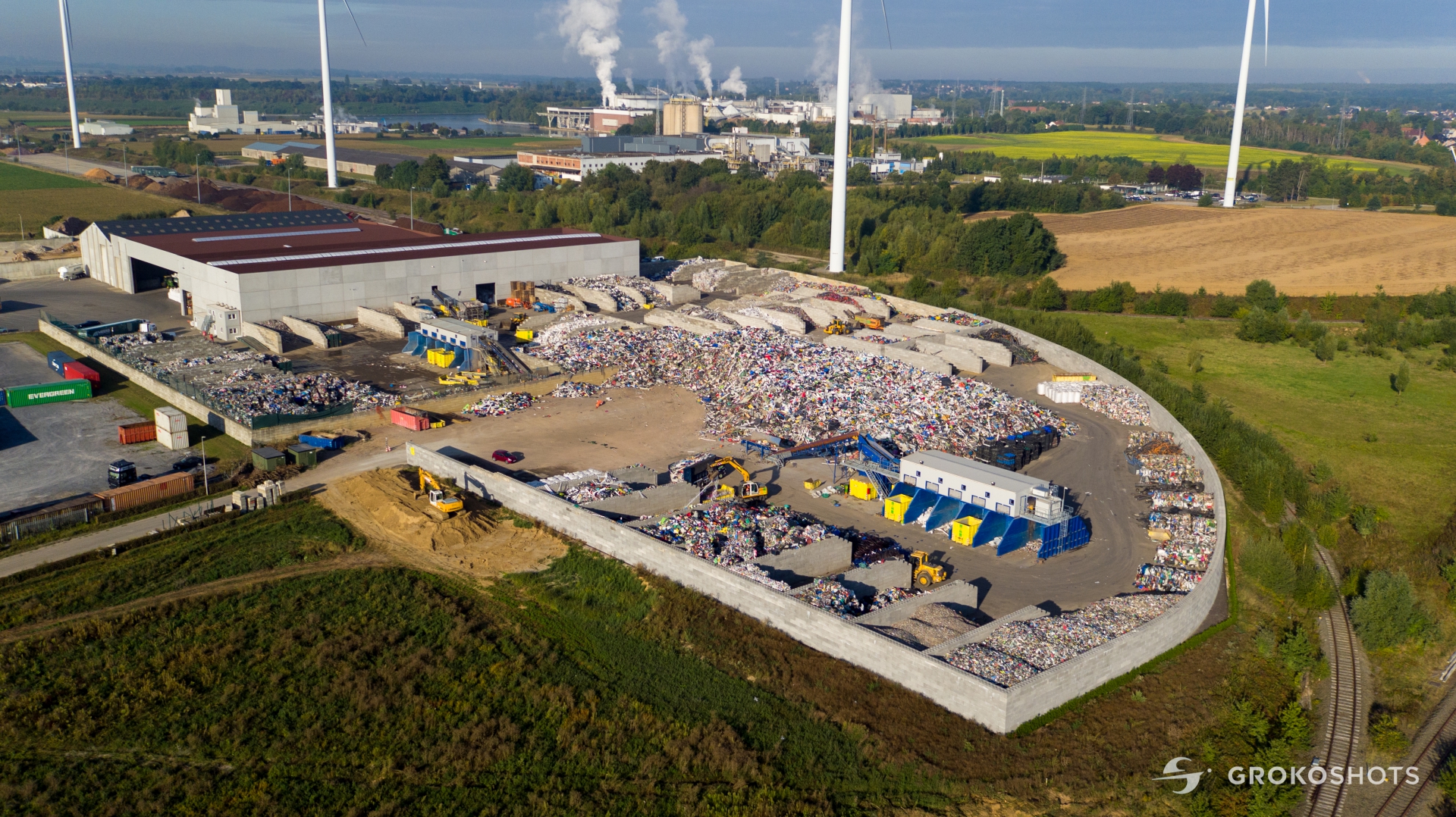 Luchtfoto van de recyclinglocatie met grote bergen gesorteerd plastic in betonnen vakken, omringd door windmolens