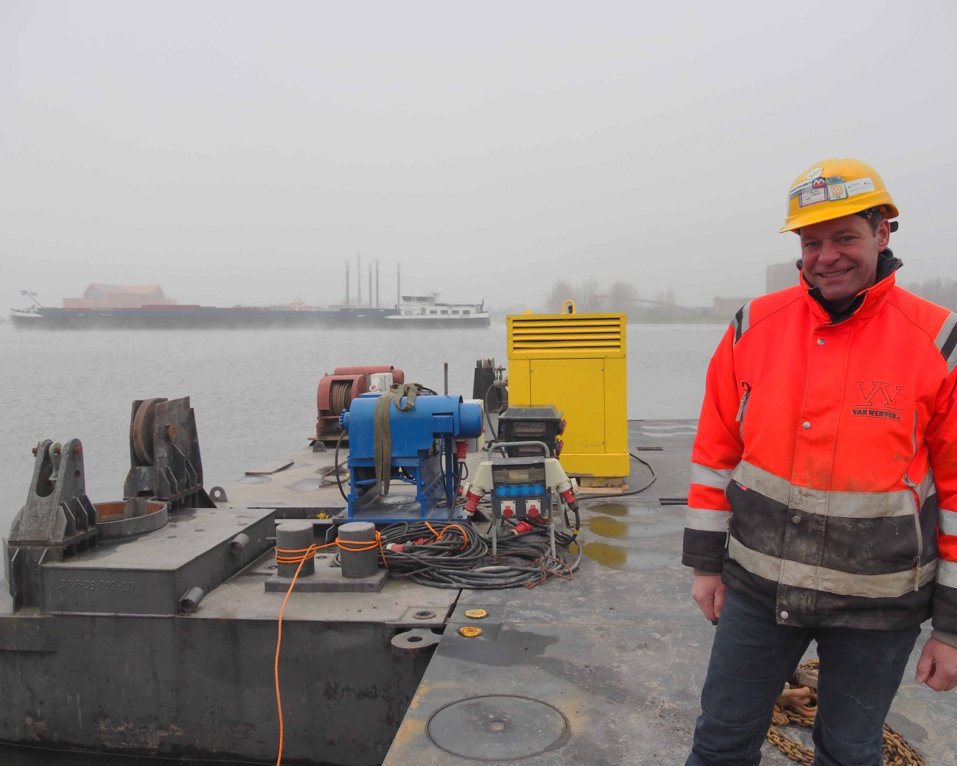 De uitvoerder staat lachend in oranje Van Werven-jas en met gele helm op het ponton, met een binnenvaartschip in de mist erachter