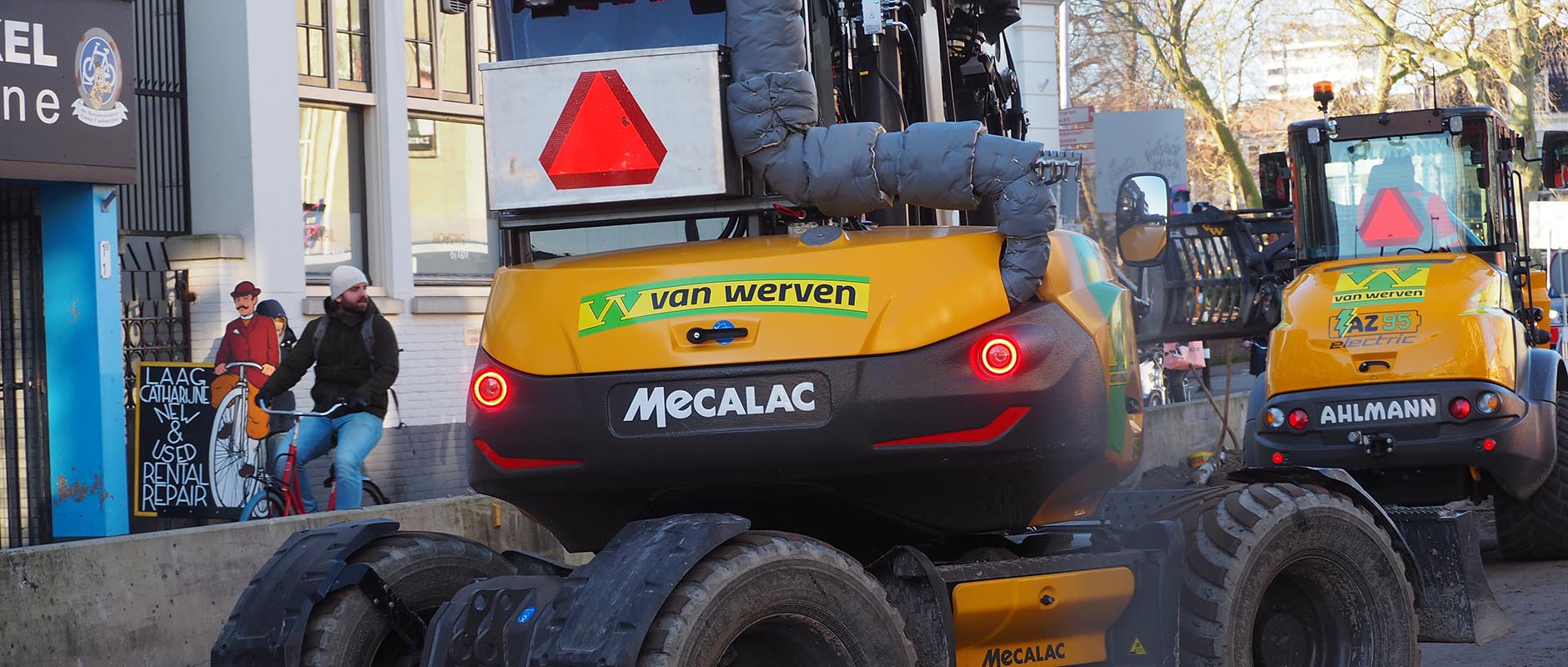 De elektrische Mecalac-graafmachine en Ahlmann-shovel van Van Werven in de straat, terwijl fietsers langs het werk rijden