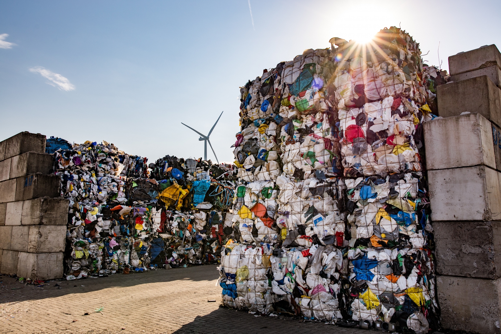 Balen geperst hard plastic tussen betonblokken op het terrein, met een windmolen en tegenlicht van de zon