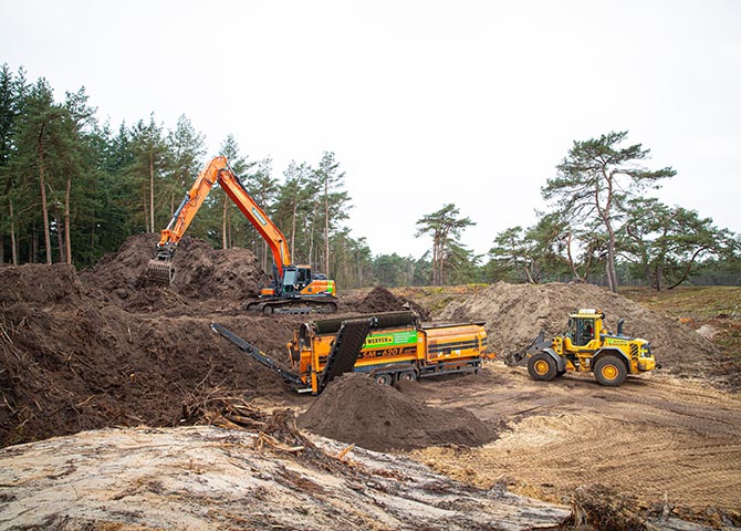 De trommelzeef van Van Werven scheidt de bosgrond, gevoed door de Doosan; een Volvo-shovel werkt aan de berg gezeefd zand