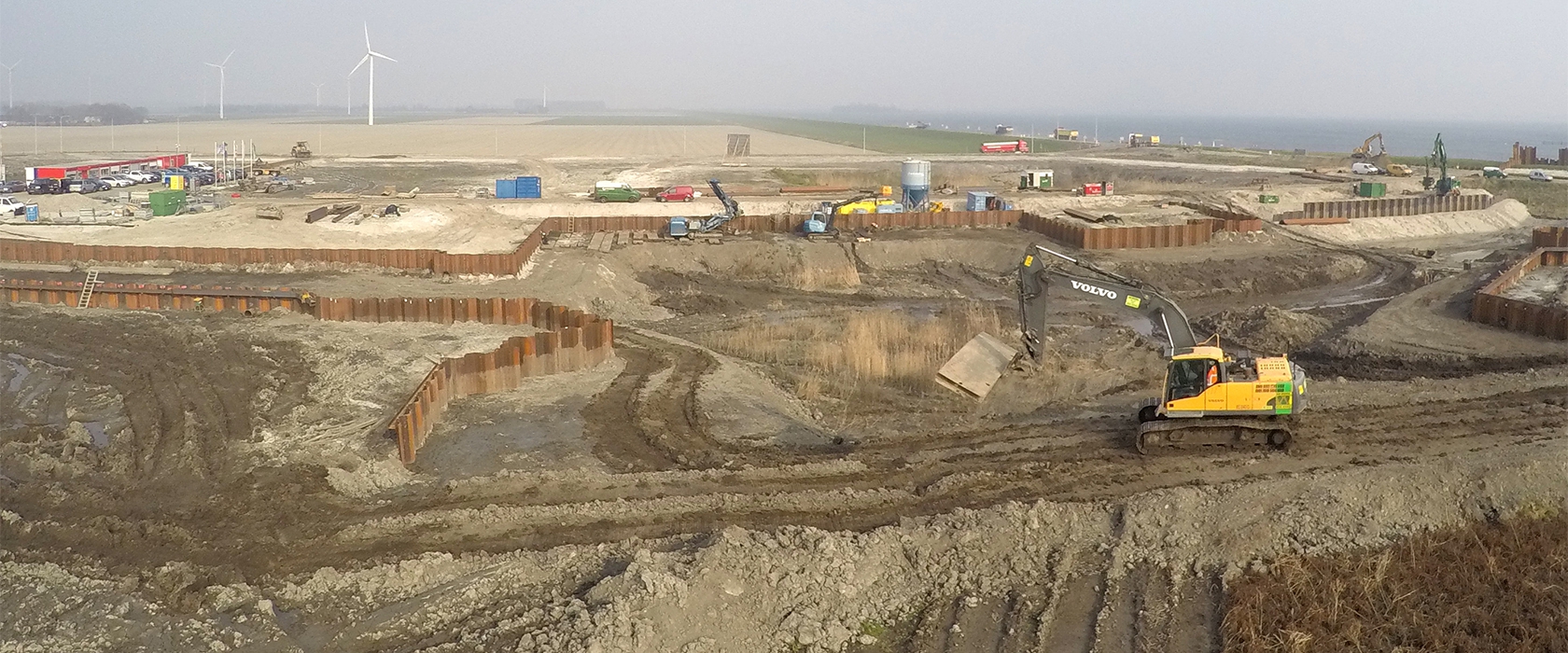 Overzicht van het werkterrein aan het water: damwandkuipen, een Volvo-rupskraan en bouwketen, met windmolens in de verte