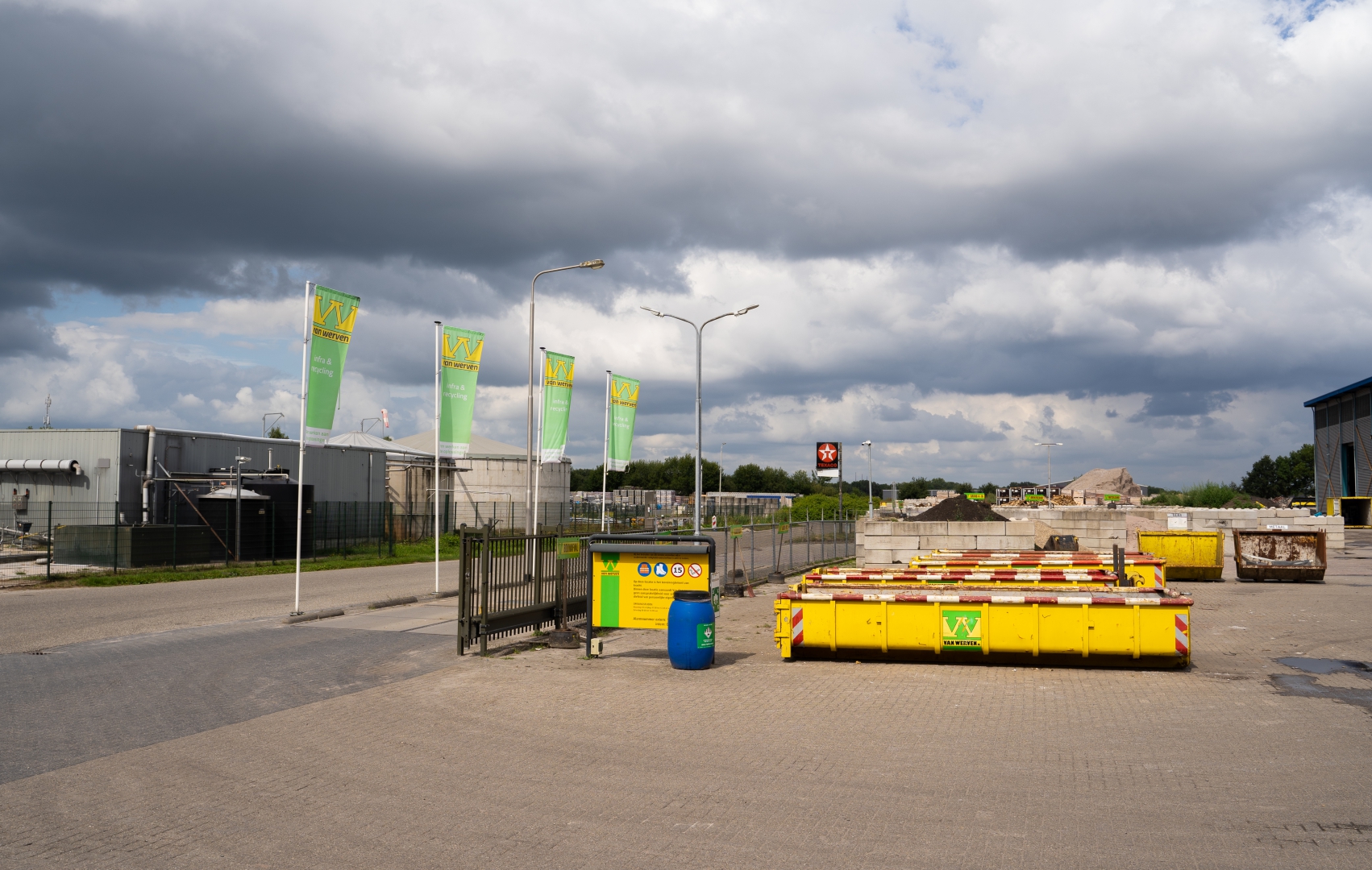 Ingang van de recyclinglocatie met vier Van Werven-vlaggen, een informatiebord en gestapelde gele containers onder een dreigende lucht