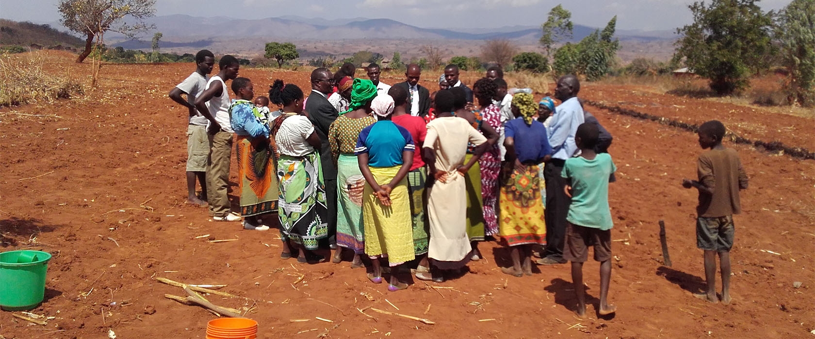 Dorpsbewoners in kleurige kleding staan in een kring op de rode akkergrond, met de bergen van Malawi op de achtergrond