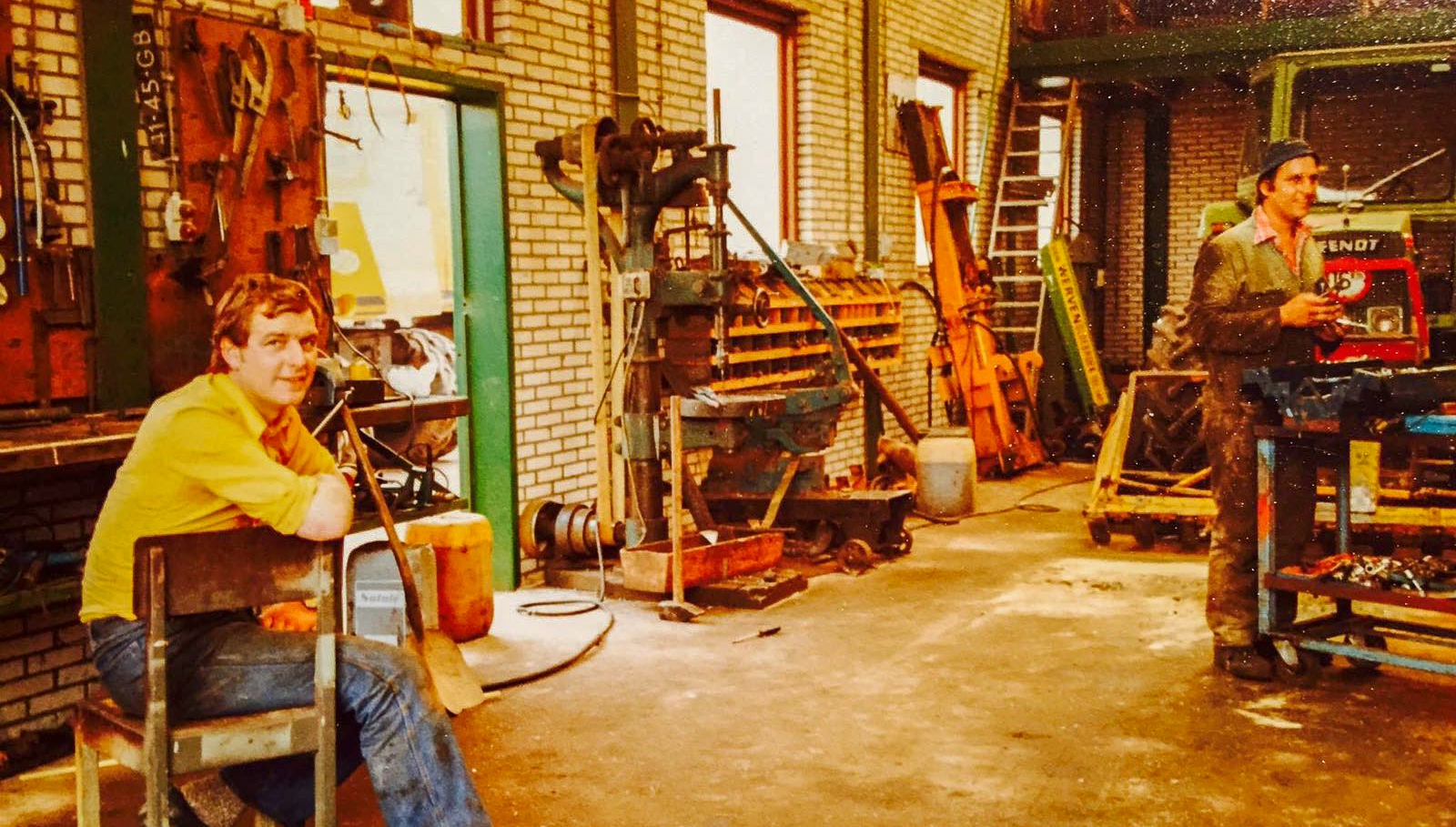 De werkplaats in de jaren tachtig: een jonge medewerker in geel overhemd op een kruk, een collega aan de onderdelenkar bij een Fendt