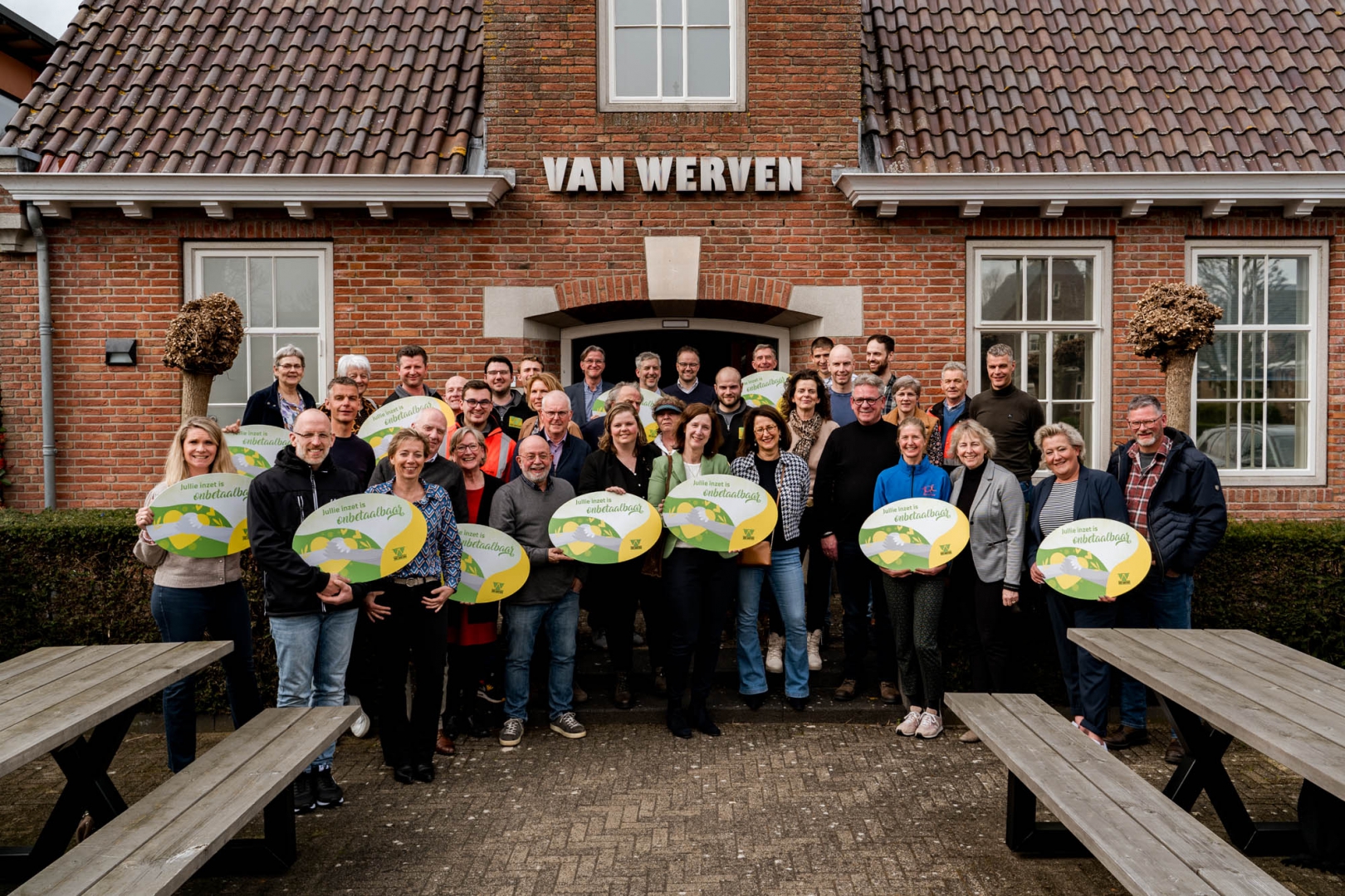 Groepsfoto van vertegenwoordigers van veertien goede doelen met ronde borden 'Jullie inzet is onbetaalbaar' voor het Van Werven-kantoor