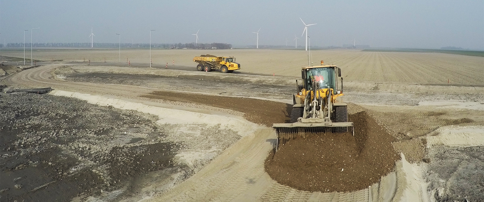 Een Volvo-shovel spreidt granulaat over het cunet van de nieuwe weg terwijl een dumper af en aan rijdt; aan de horizon draaien windmolens
