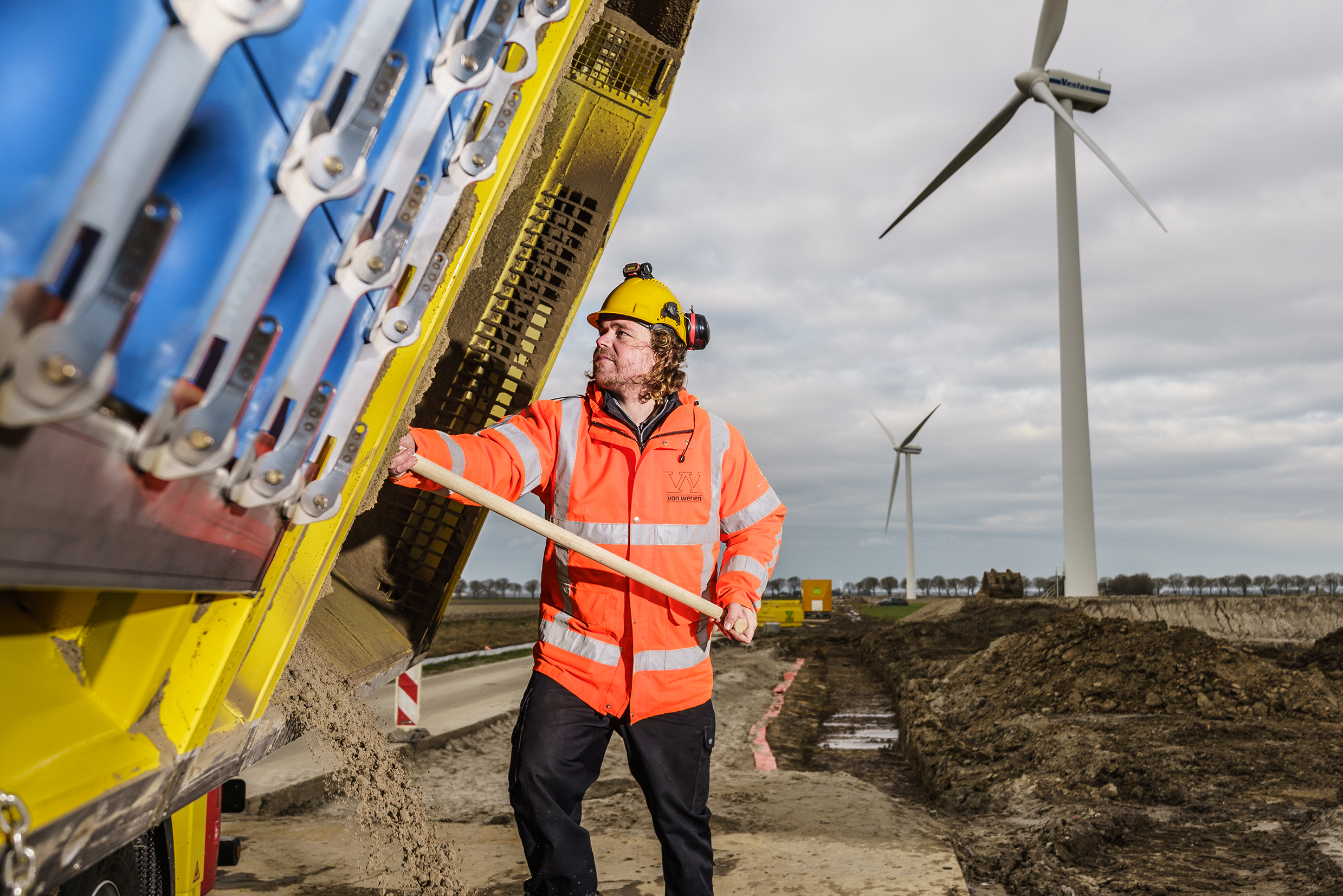 Een medewerker in oranje jas werkt bij de gekiepte laadbak van de zuigwagen, met windmolens op de achtergrond